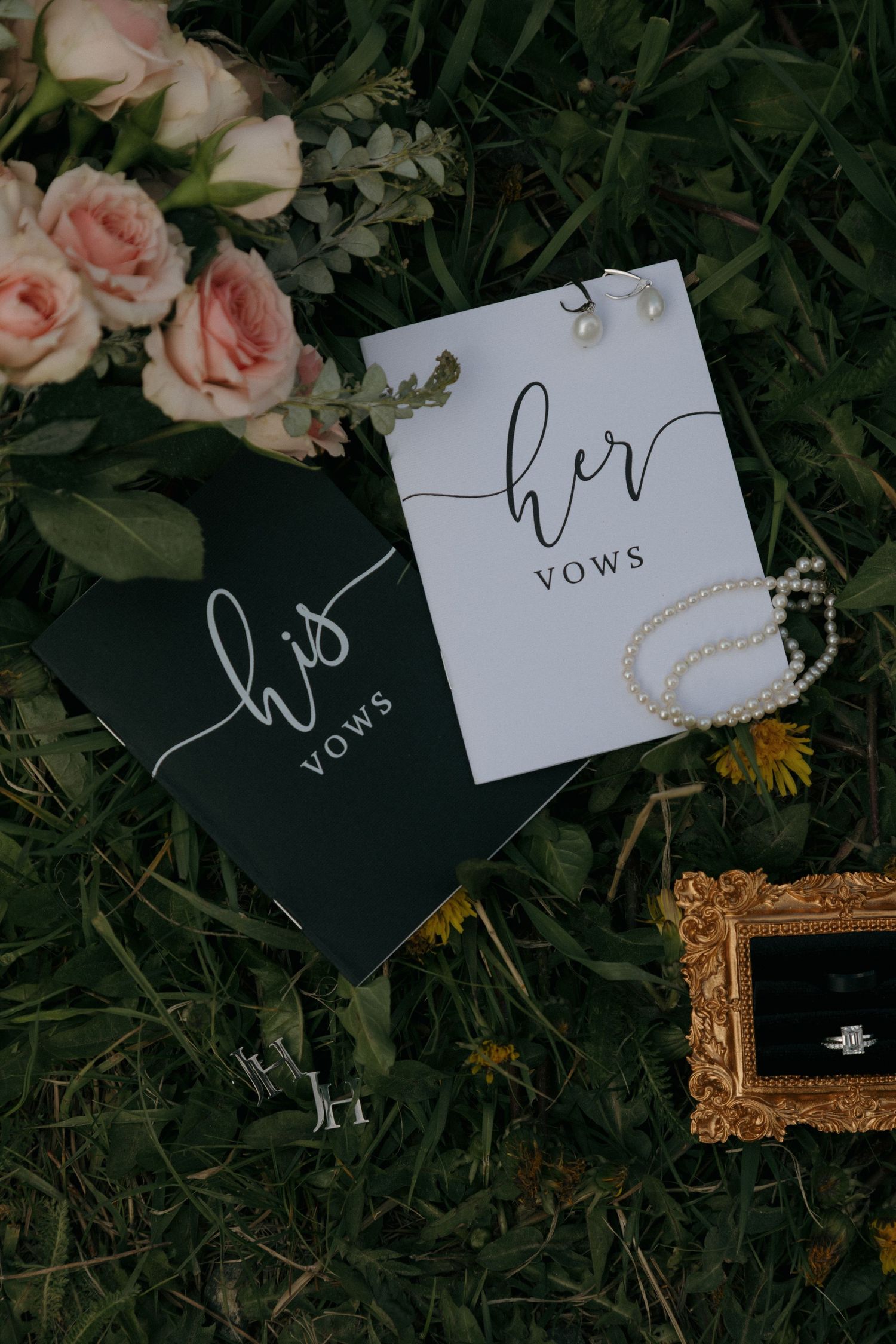 His and her vow books with pearl bracelet and ring in gold frame on green grass with pink roses.