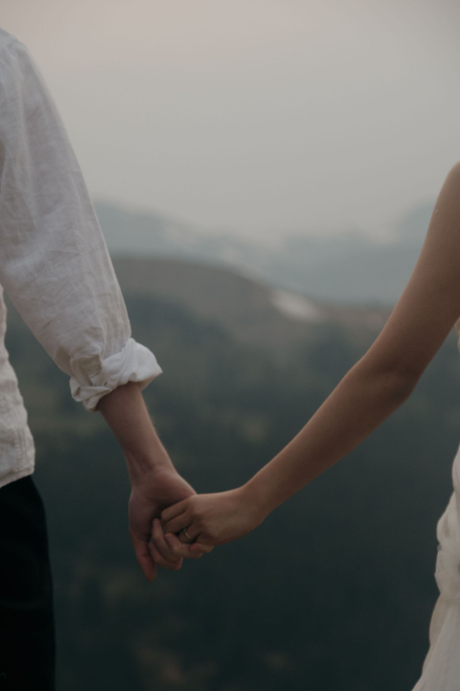 Close-up of bride and groom holding hands against a soft misty mountain backdrop during their outdoor wedding.