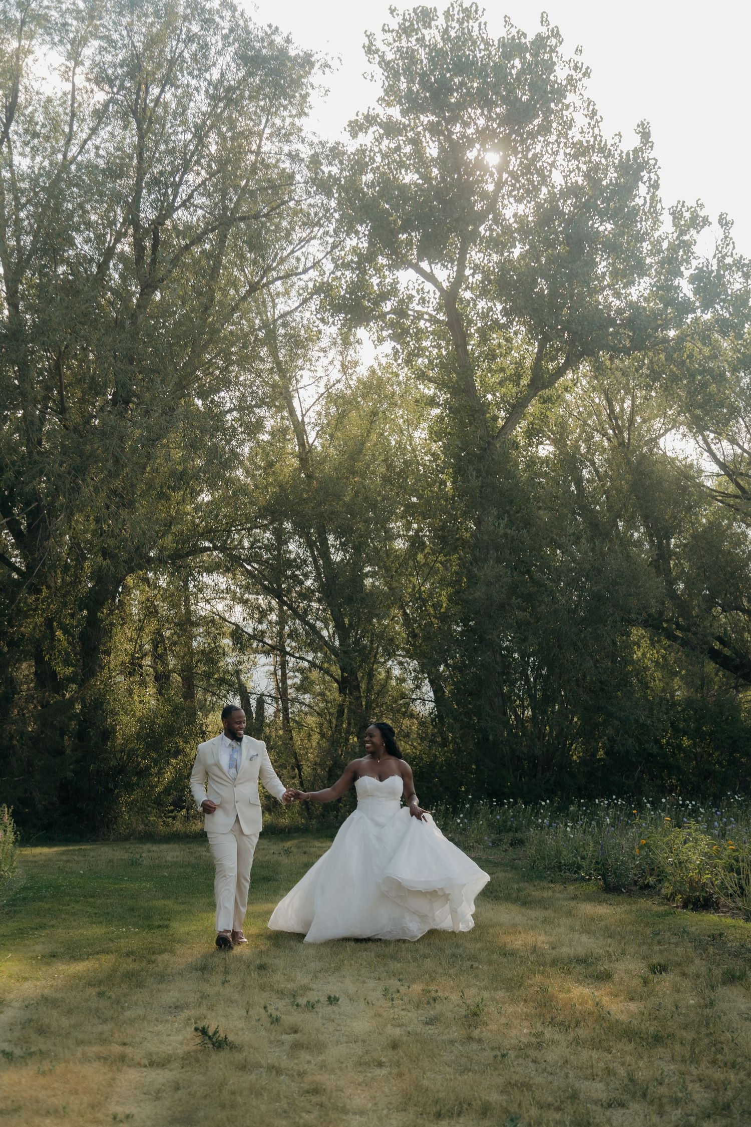 Black couple in wedding attire holding hands and walking through a sunlit green meadow surrounded by tall trees.