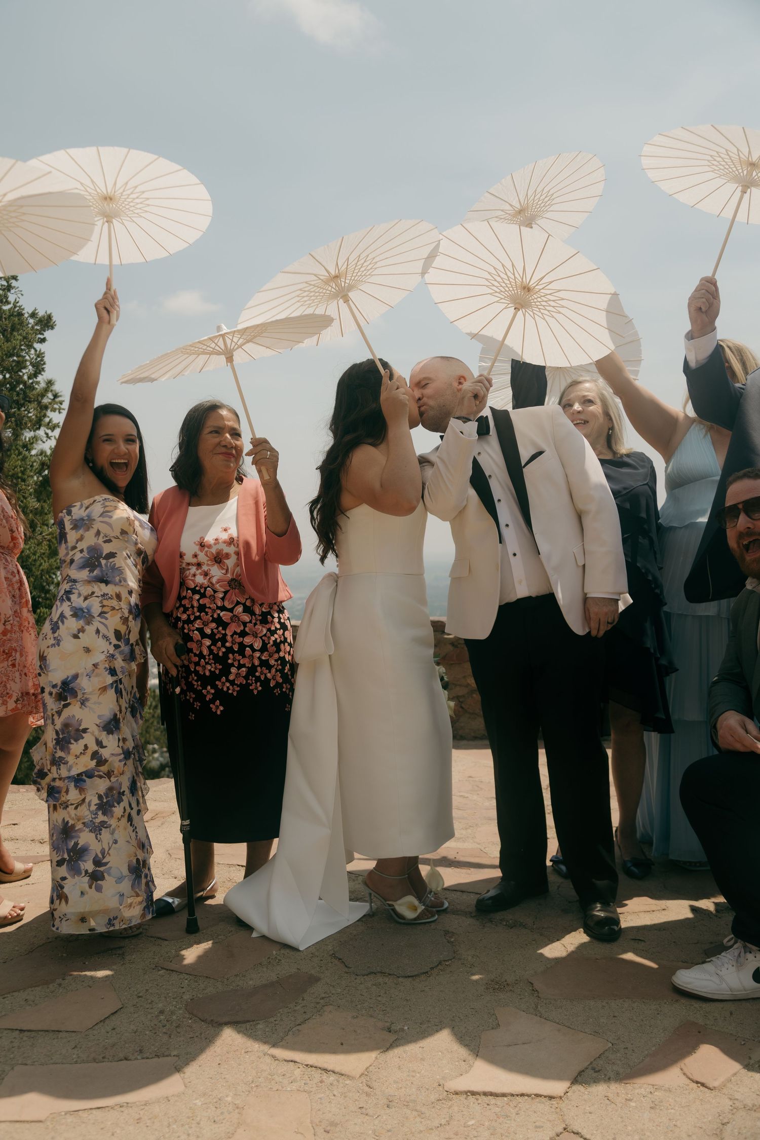Newlyweds kiss surrounded by joyful guests holding white parasols aloft at an outdoor wedding celebration on a sunny day.