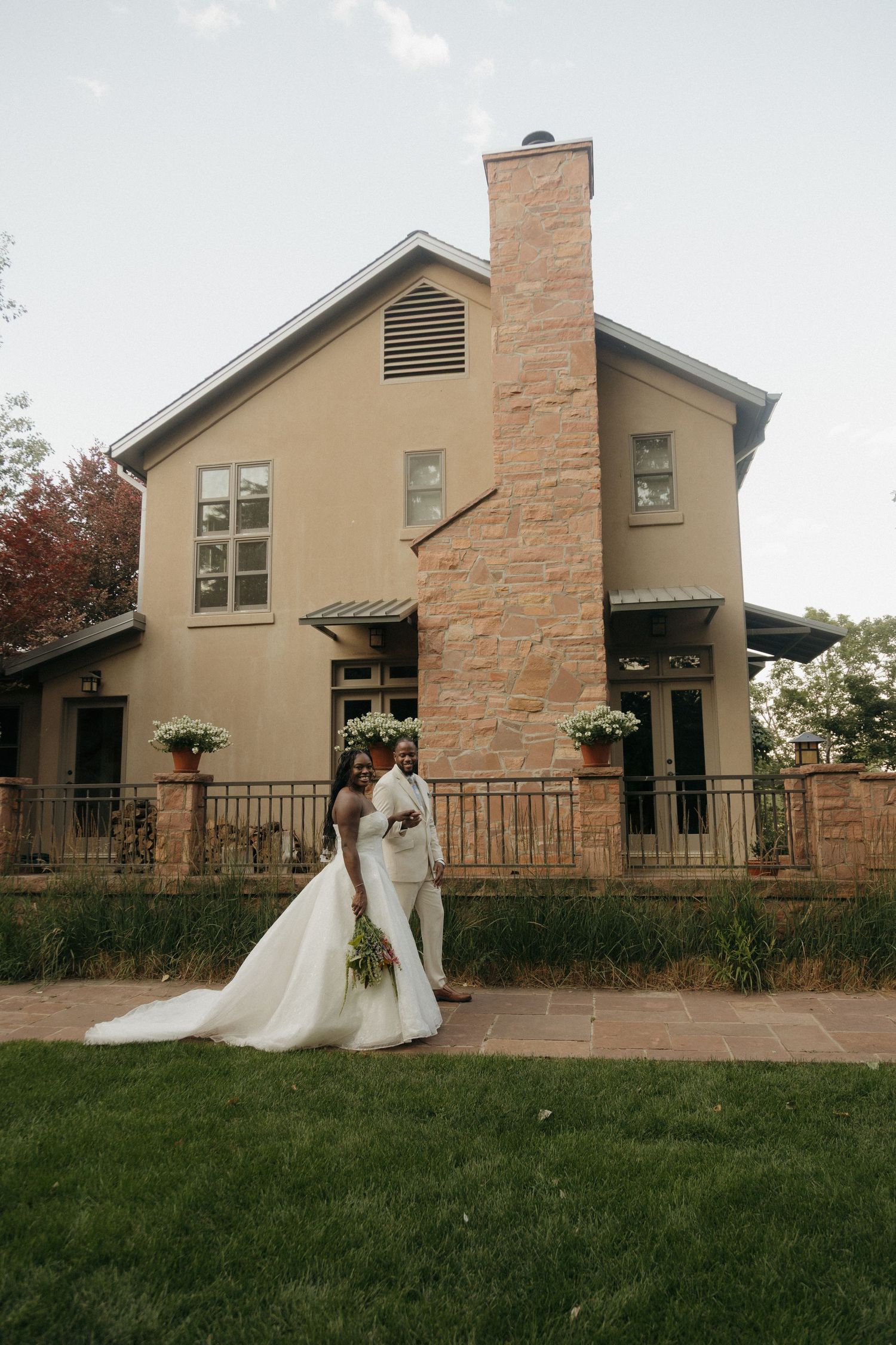 Black bride and groom pose in front of an elegant stucco and stone venue building with a tall chimney.