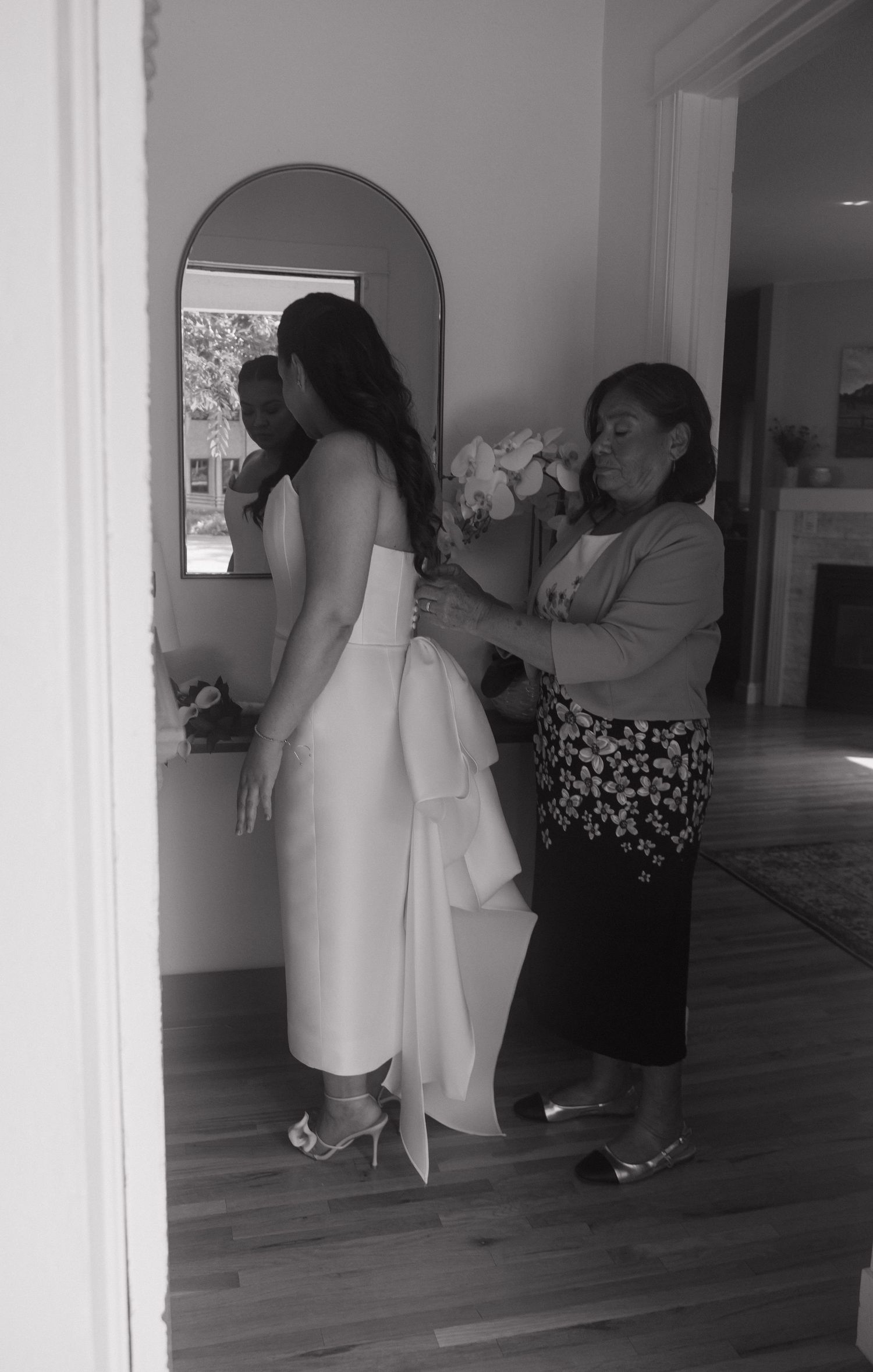 Black and white photo of a woman helping a bride in a white dress prepare near a mirror before her wedding.