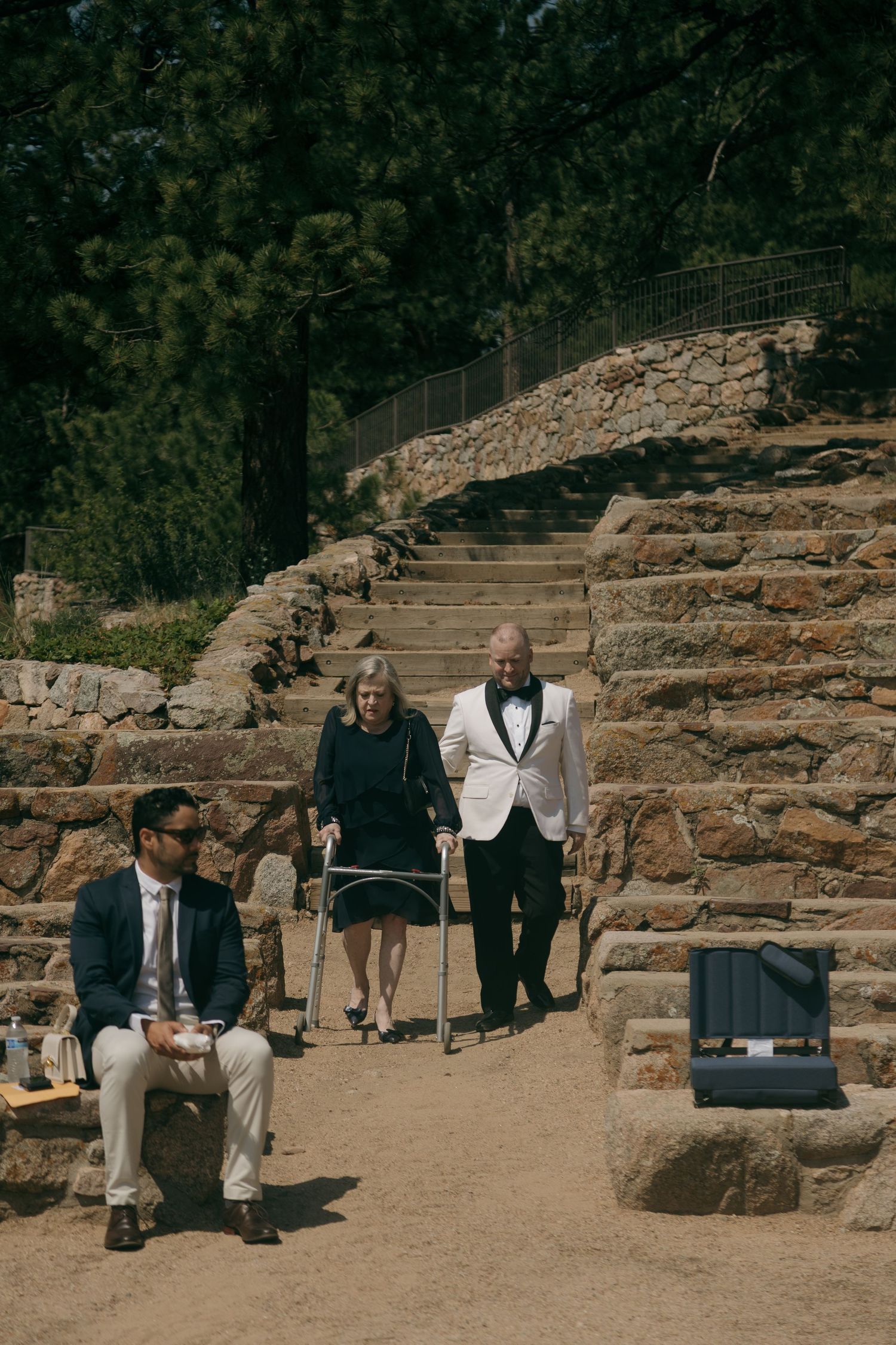 Groom in white tuxedo escorts elderly woman with walker down stone steps at outdoor wedding venue.