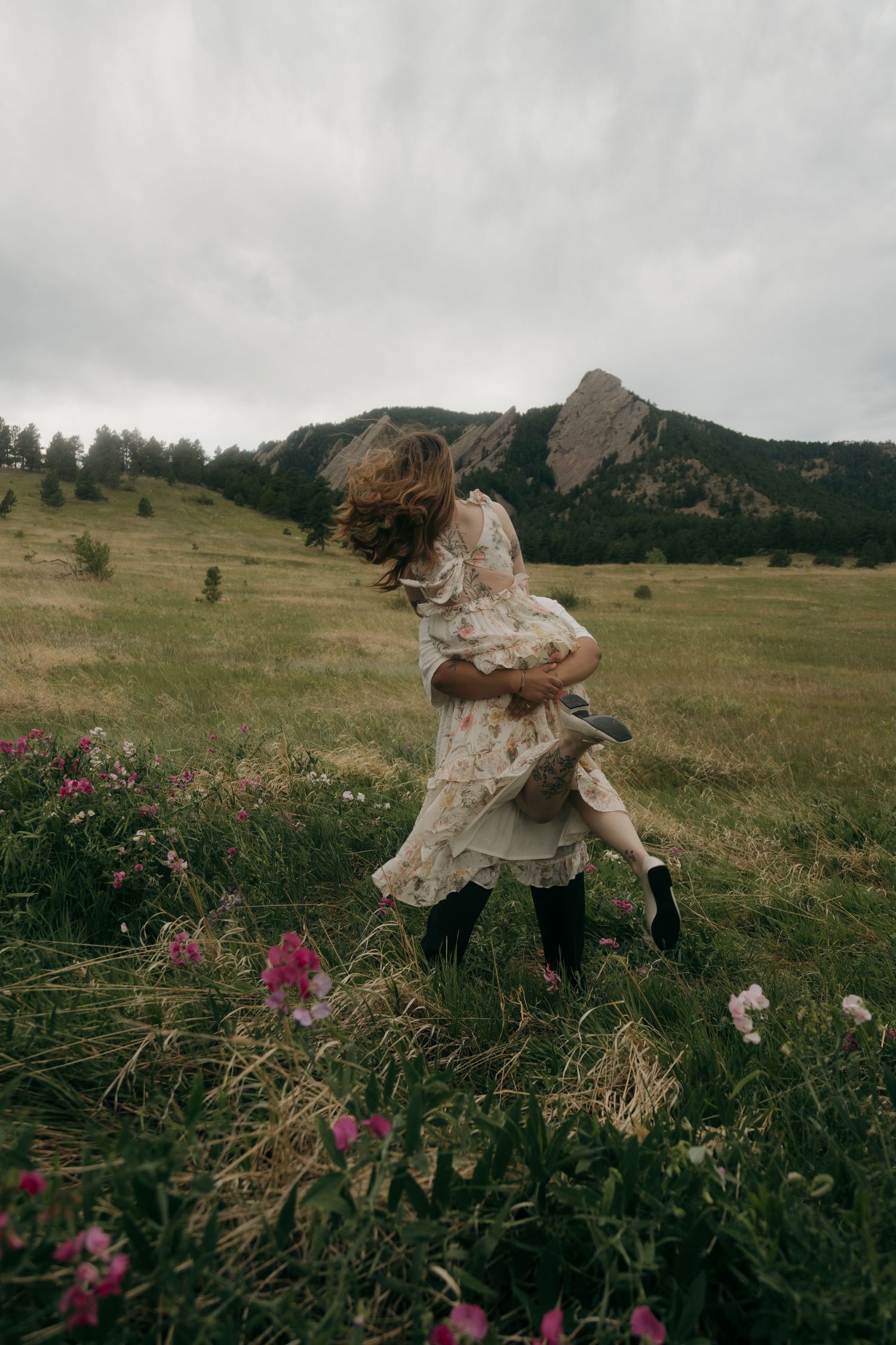 Woman in floral dress dancing freely in a windy meadow with pink wildflowers and mountain backdrop.