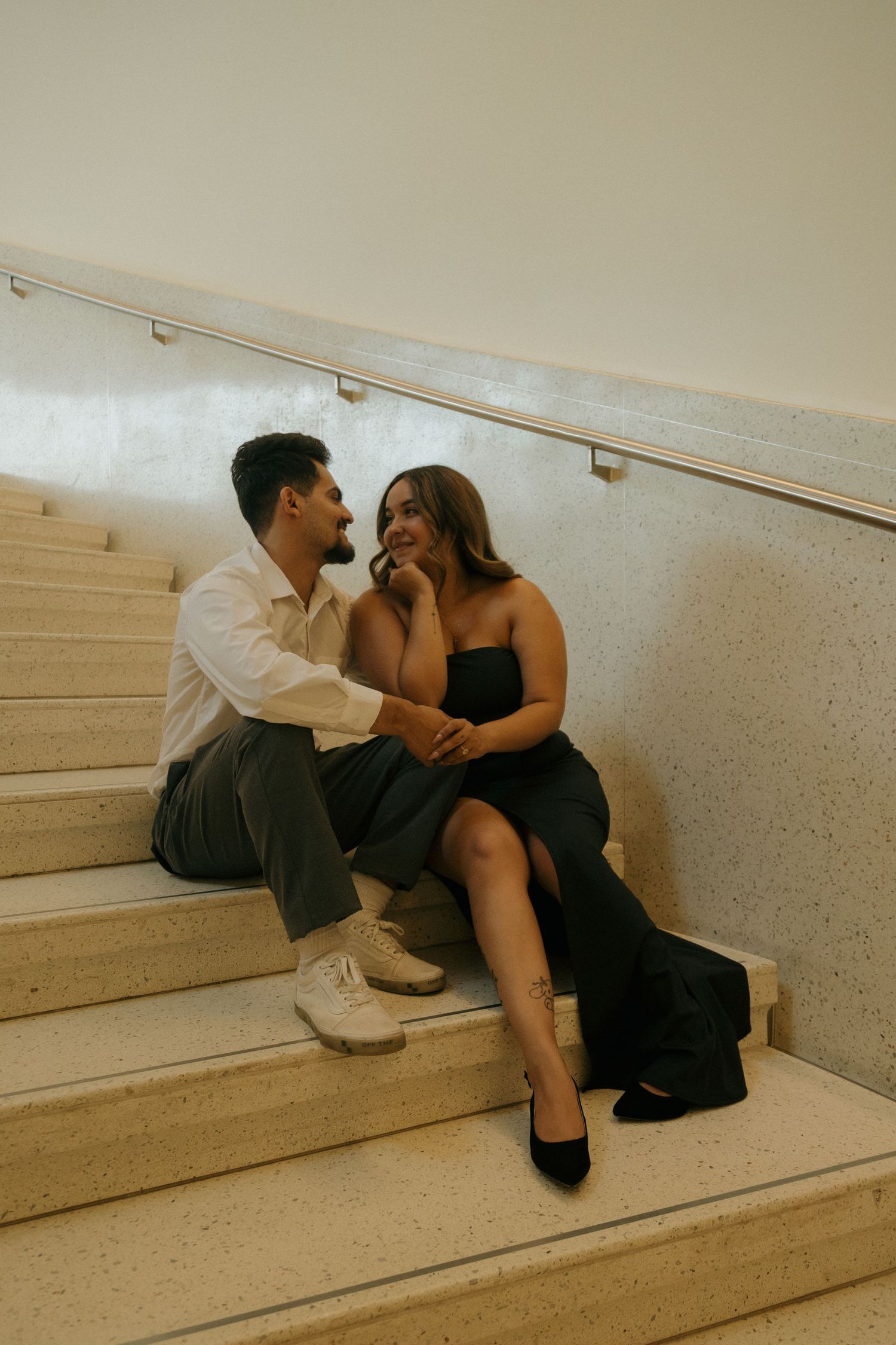 Couple sitting closely on marble stairs smiling at each other, woman in black slit dress and man in white shirt.