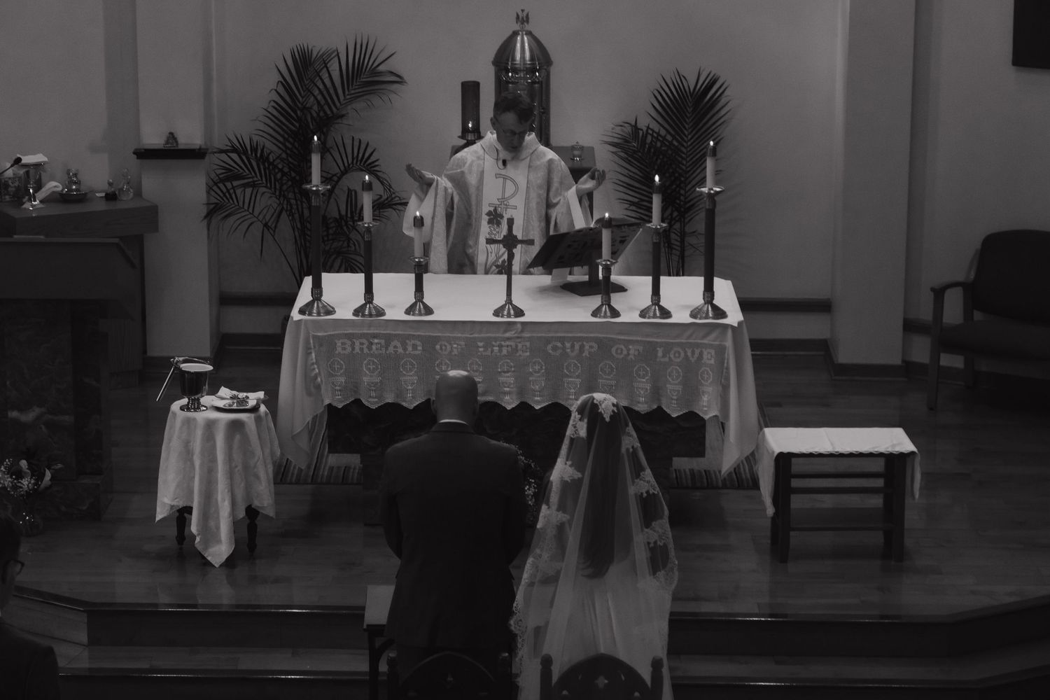 Black and white photo of a priest performing Catholic Mass at an altar with candles in a church.