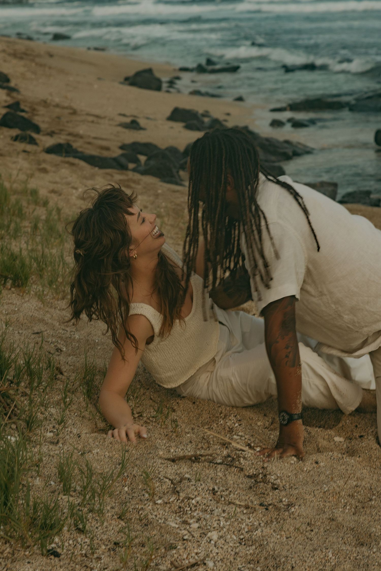 A romantic couple in beige outfits share an intimate moment on a coastal cliff overlooking the ocean.