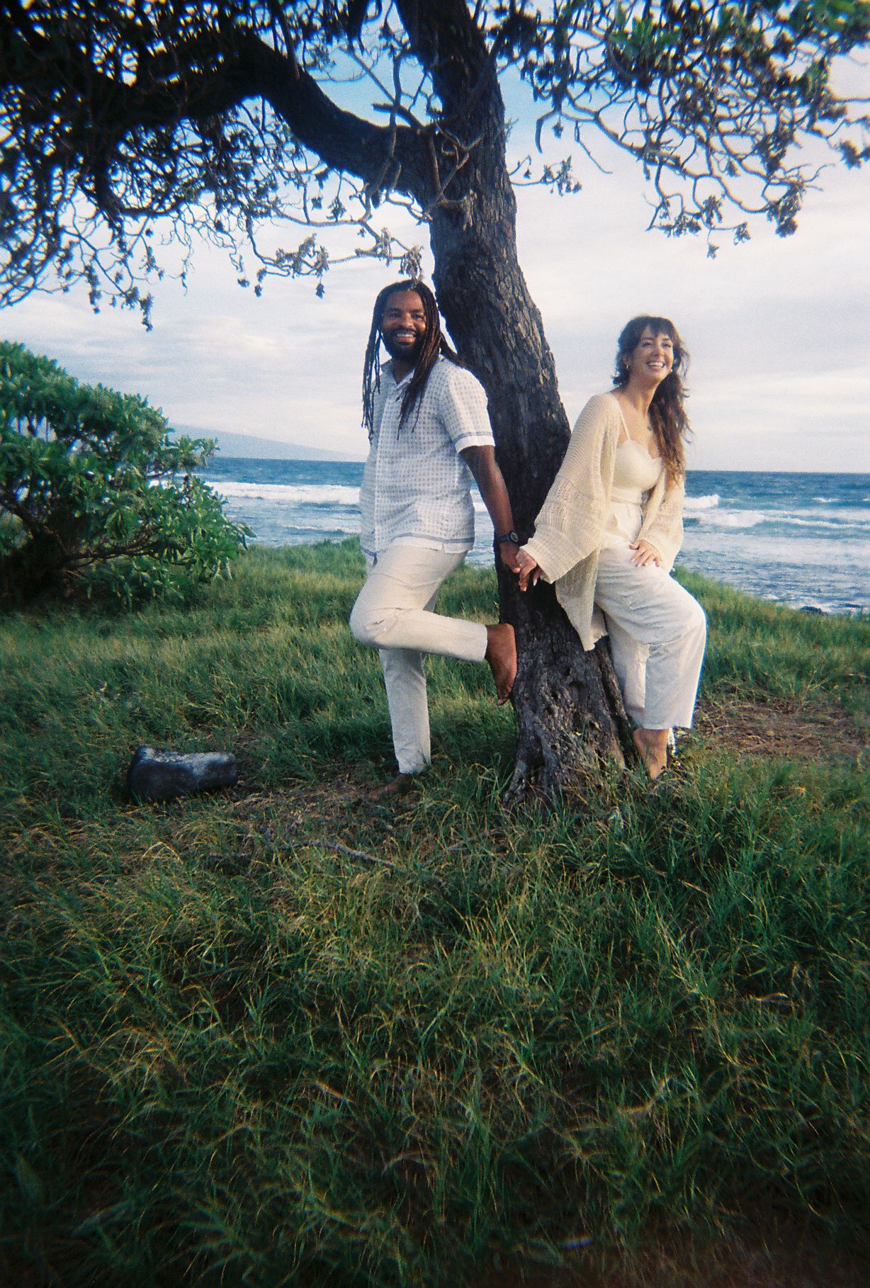 Two people in white outfits lean against a tree by the ocean on a grassy hillside at sunset.