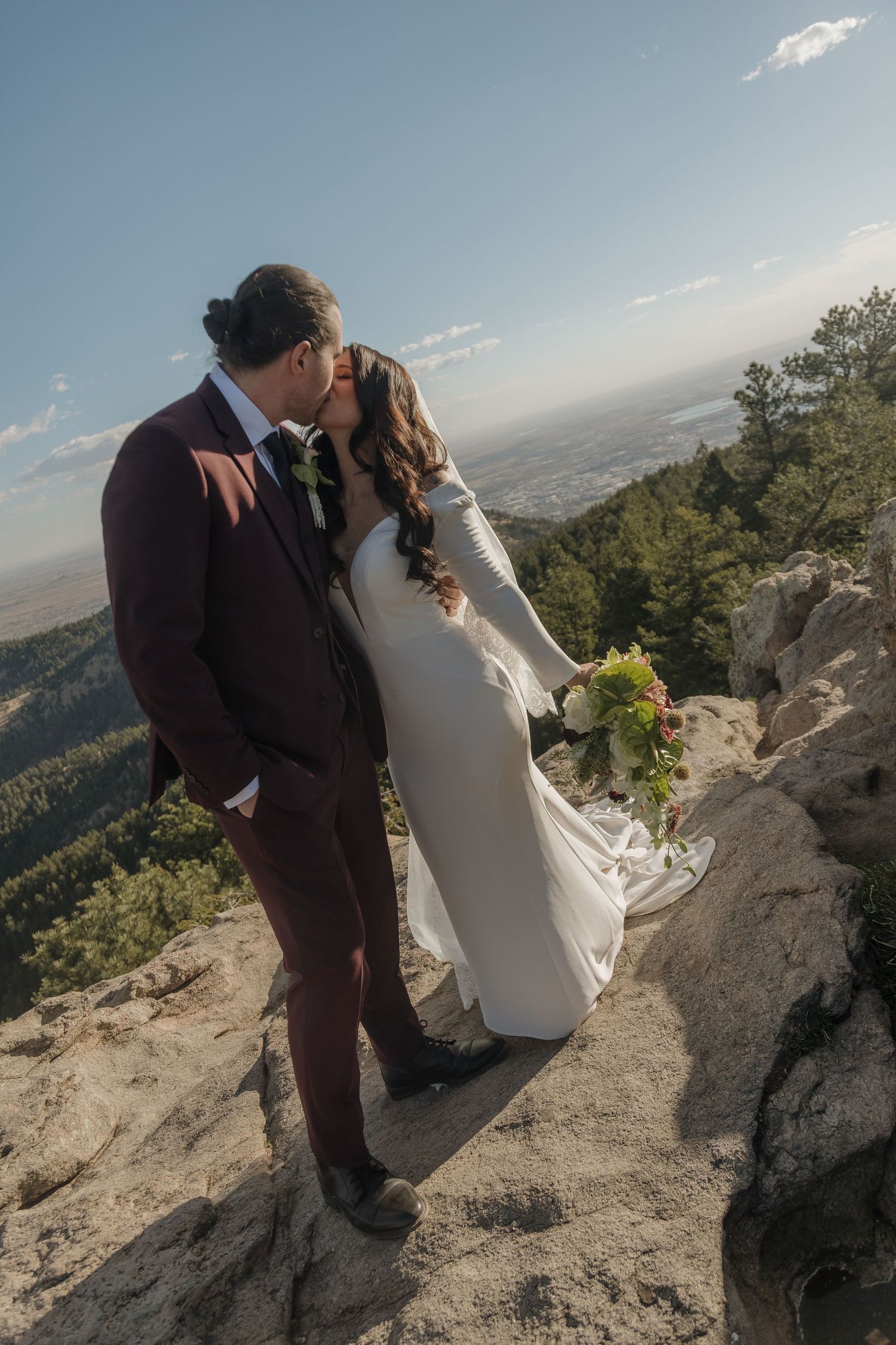 A romantic wedding couple shares a kiss on a mountaintop overlook with a scenic valley vista stretching into the distance.