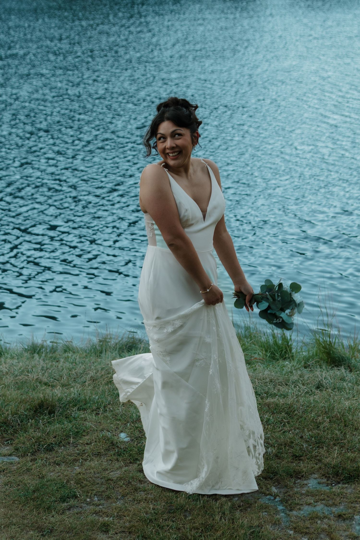 A bride in a white dress laughs joyfully by a serene lake.