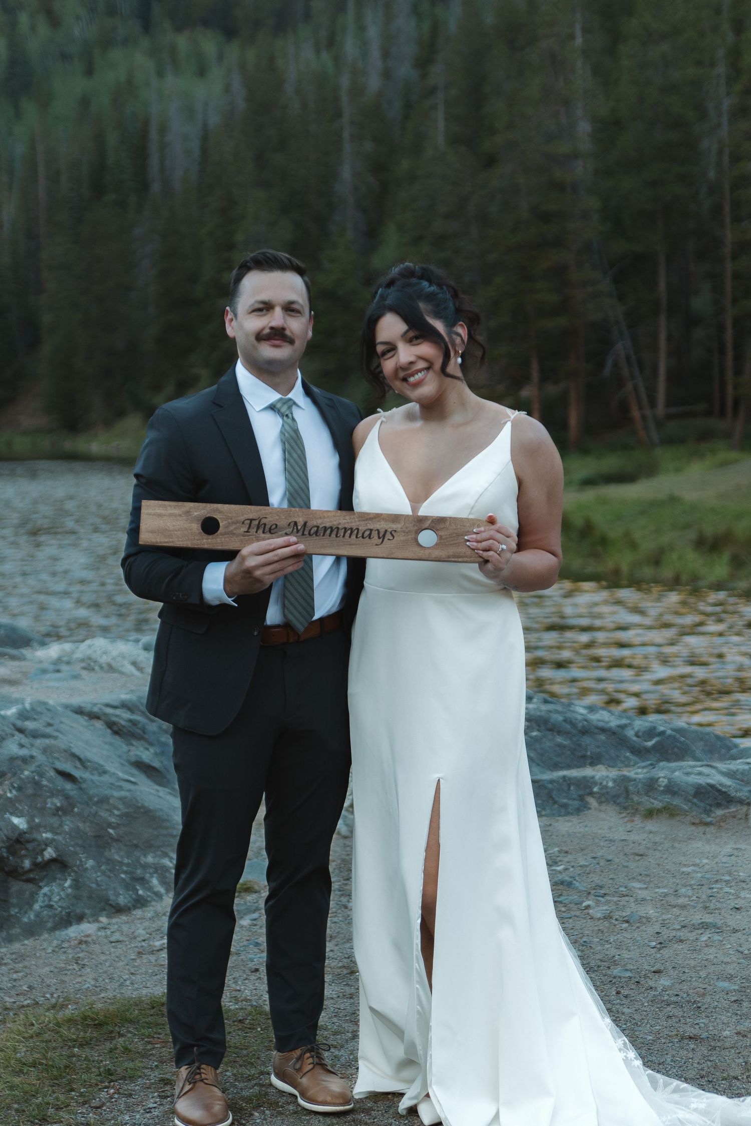 Newlyweds laughing while displaying wooden name sign at lakeside mountain venue.