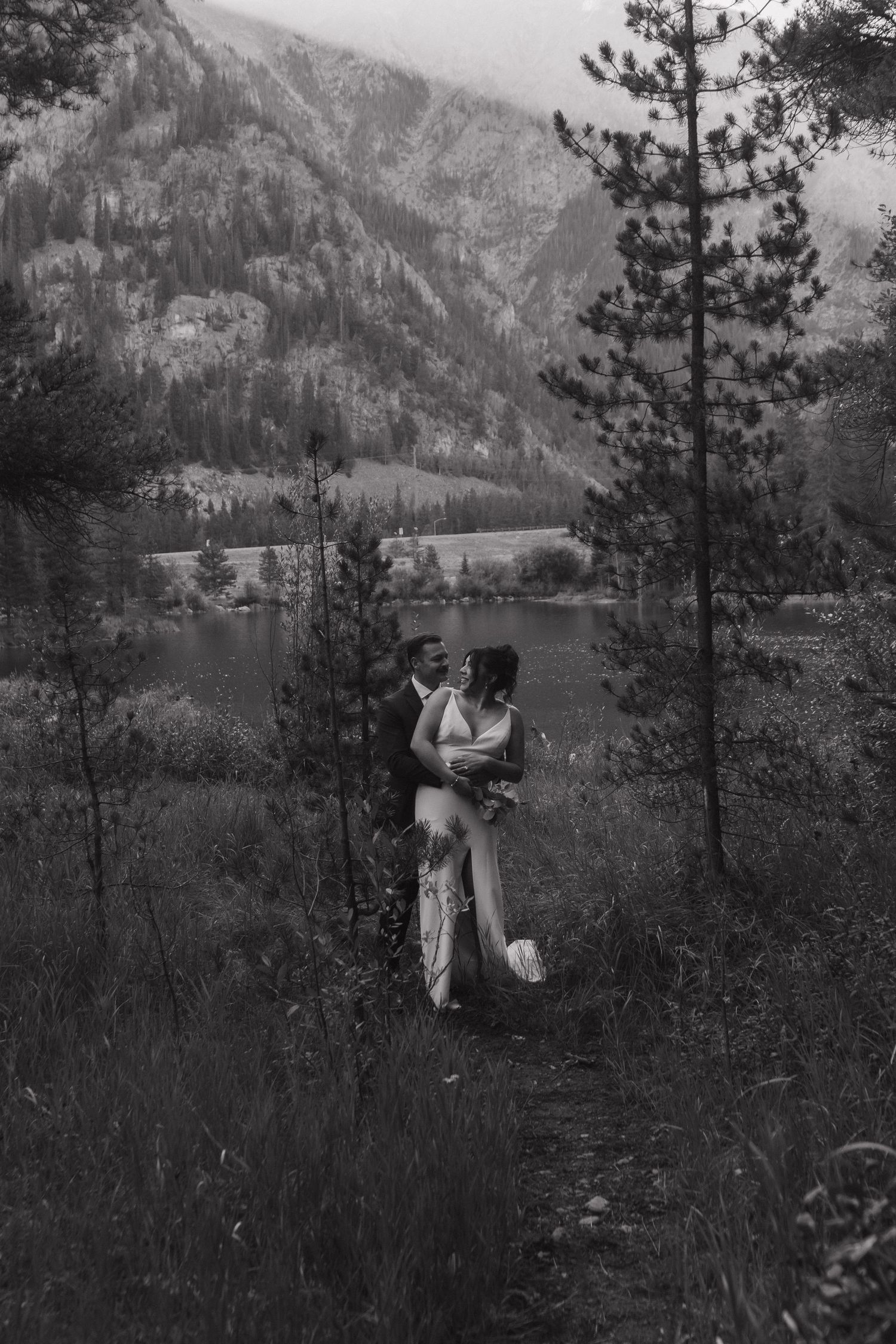 A black and white wedding photo showing an intimate moment against a mountain lake landscape.