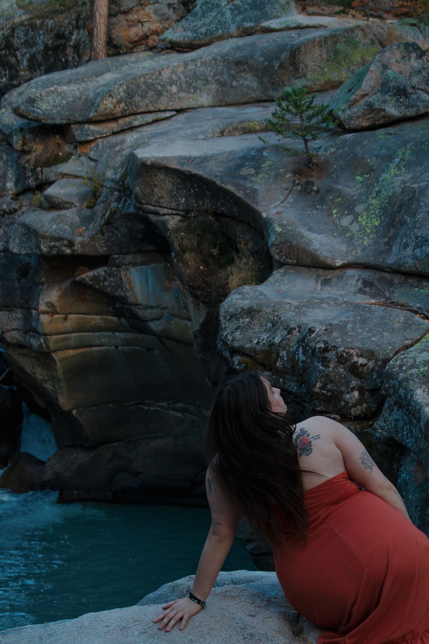 Person in a red dress sits at the edge of a rocky cliff overlooking turquoise water in a dramatic natural setting.