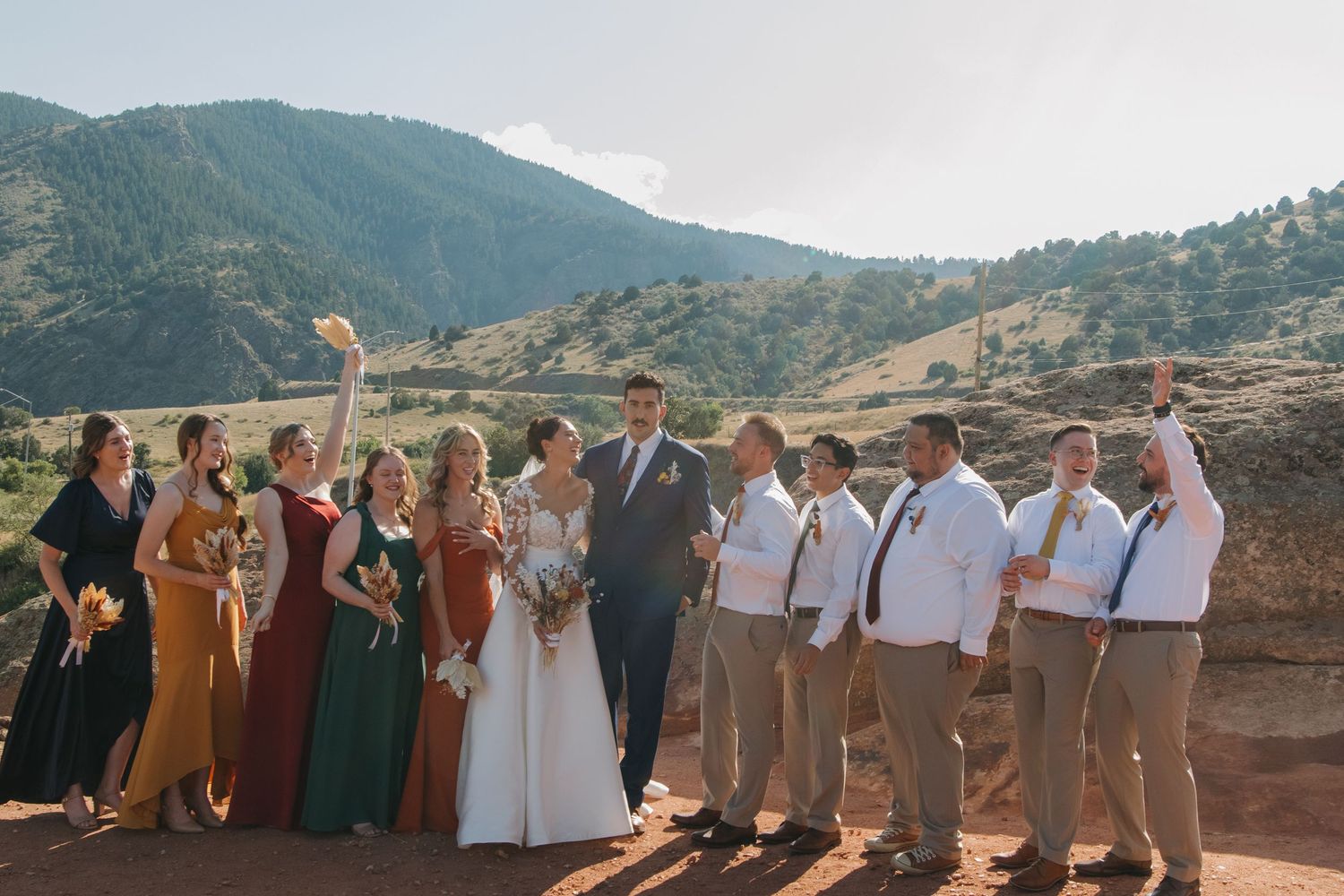 Large wedding party in mixed formal attire poses against mountain backdrop.