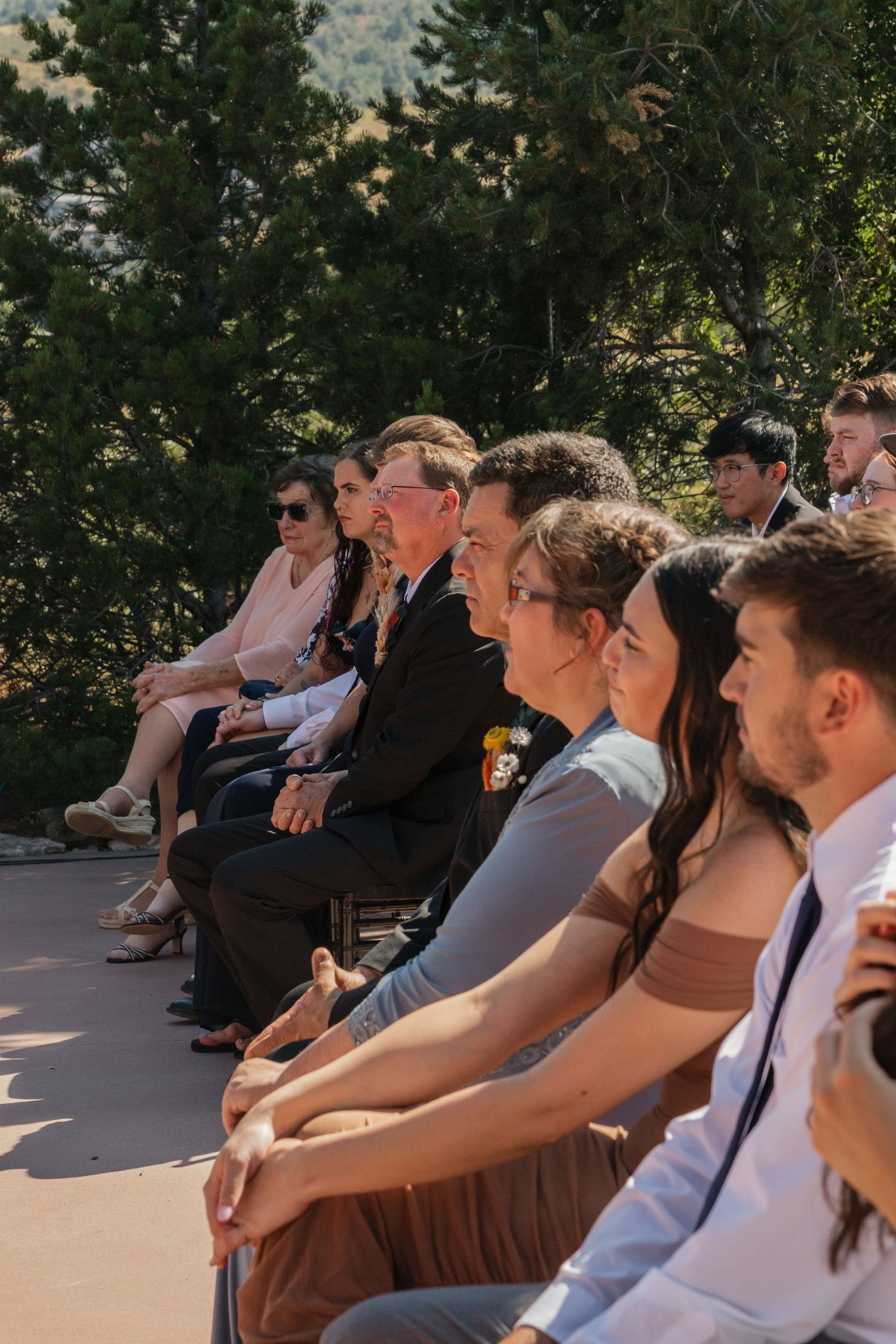 Wedding attendees seated in rows outdoors with greenery and trees in the background.