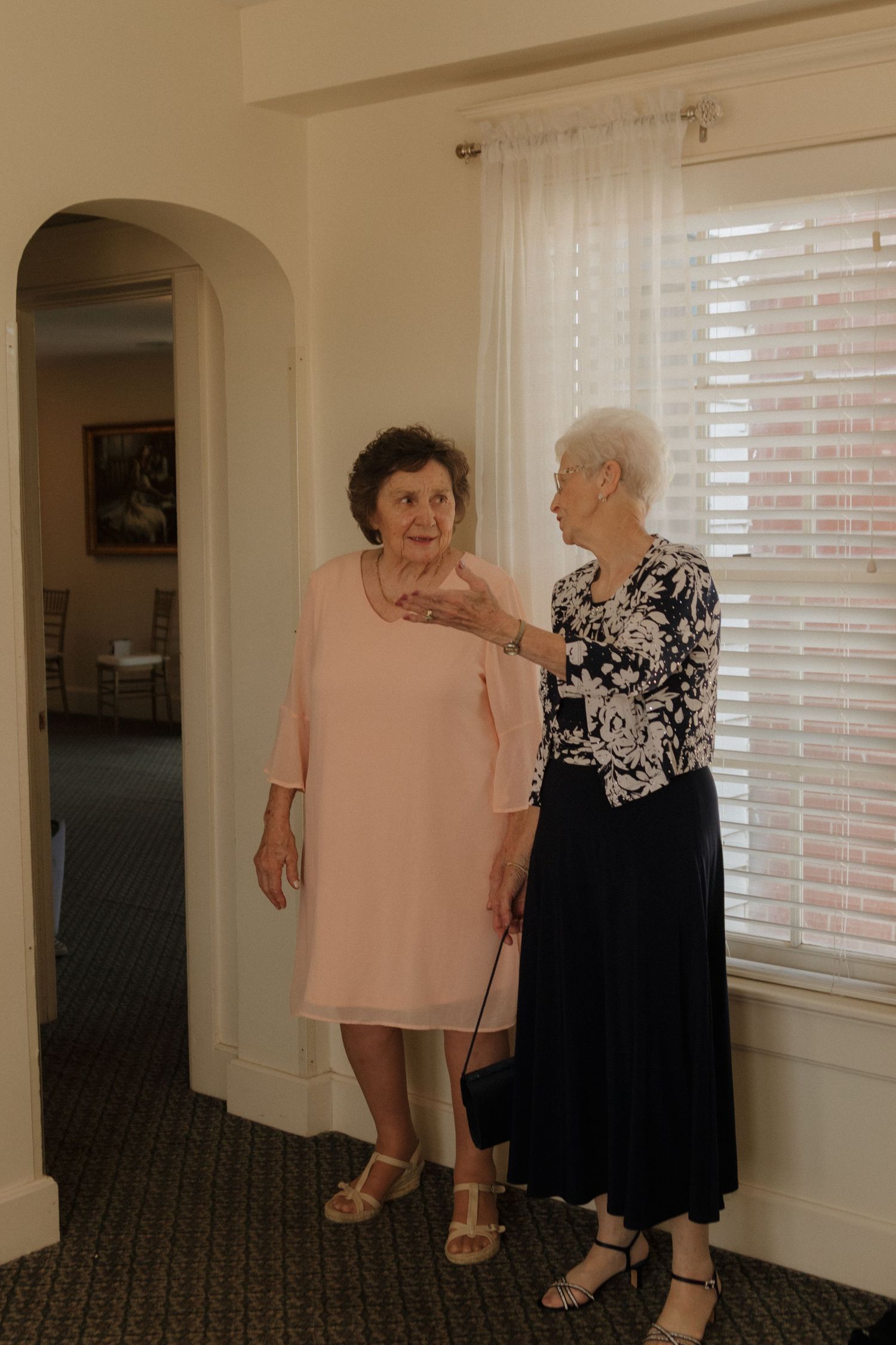 Two people in formal attire having a conversation in a brightly lit hallway with windows.