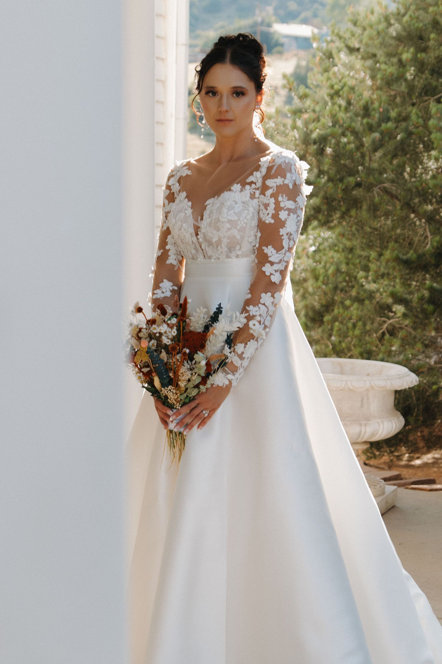 Elegant bride in long sleeve lace wedding gown holding autumn-toned bouquet.