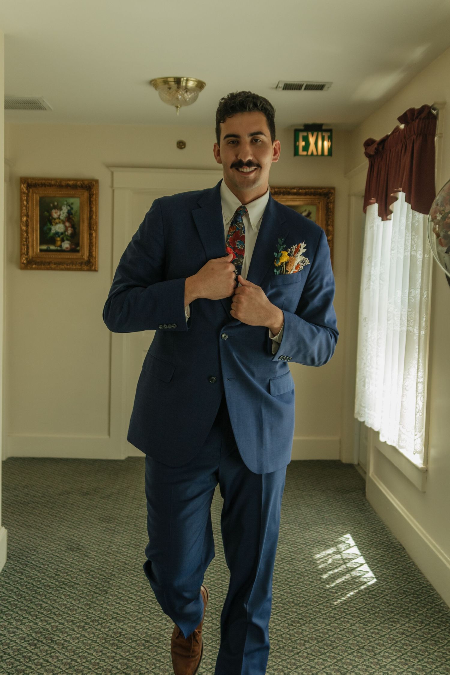 Groomsmen share candid moments while getting ready in a carpeted hallway with natural light.