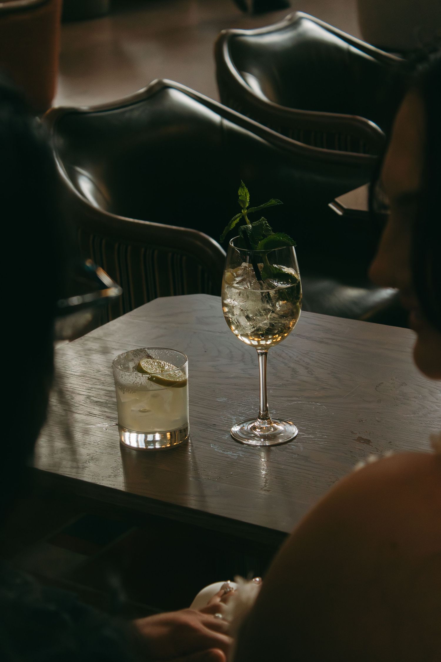 Two elegant cocktail glasses sit on a wooden table in moody lighting with dark leather seats nearby.