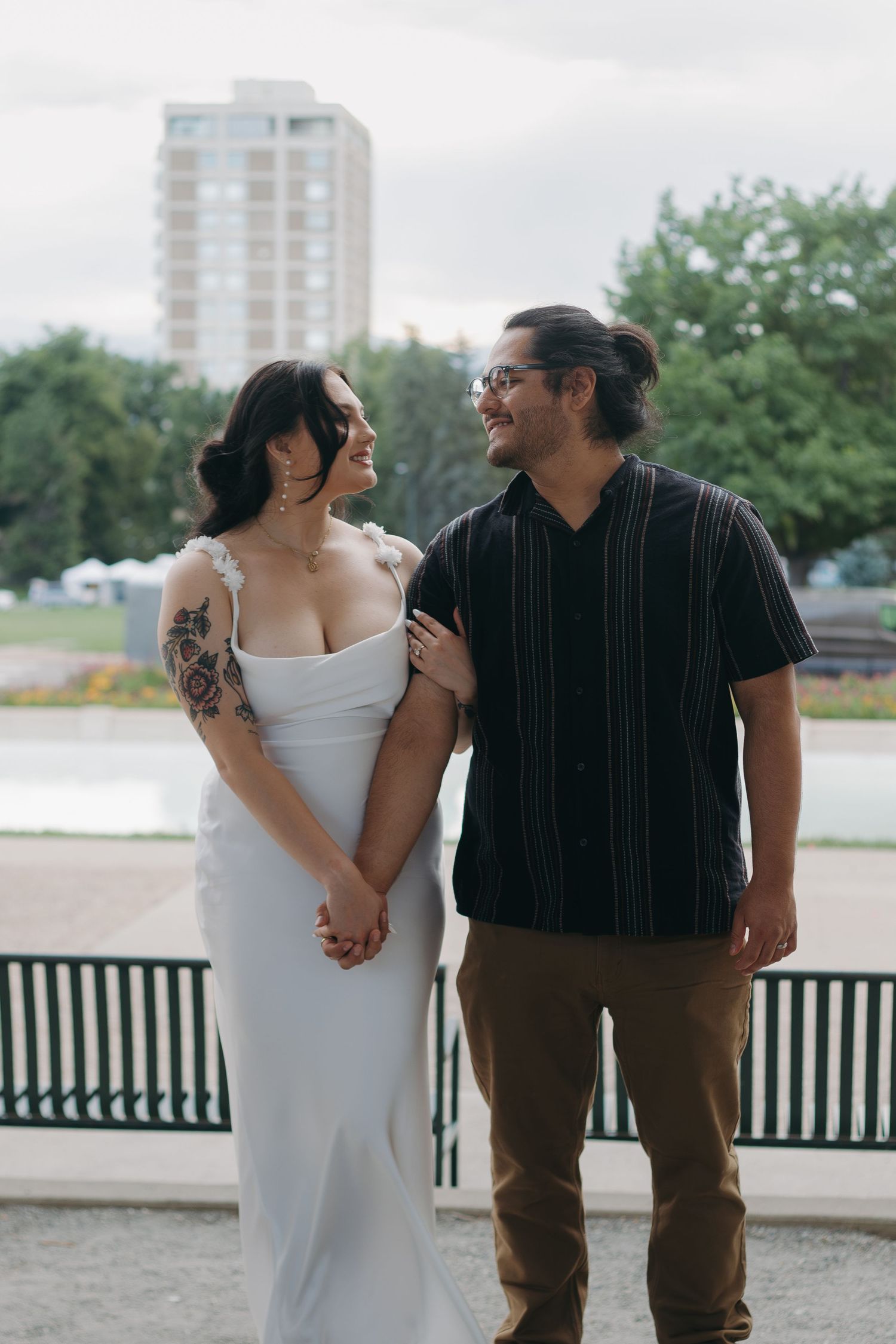 Couple poses together on waterfront promenade with city buildings in background.