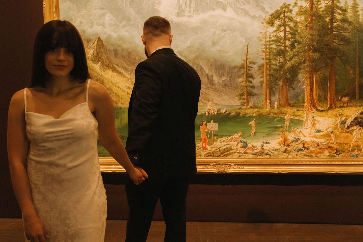 A row of isolated landscape paintings with warm earth tones hang in a gallery space with figures viewing the artwork.