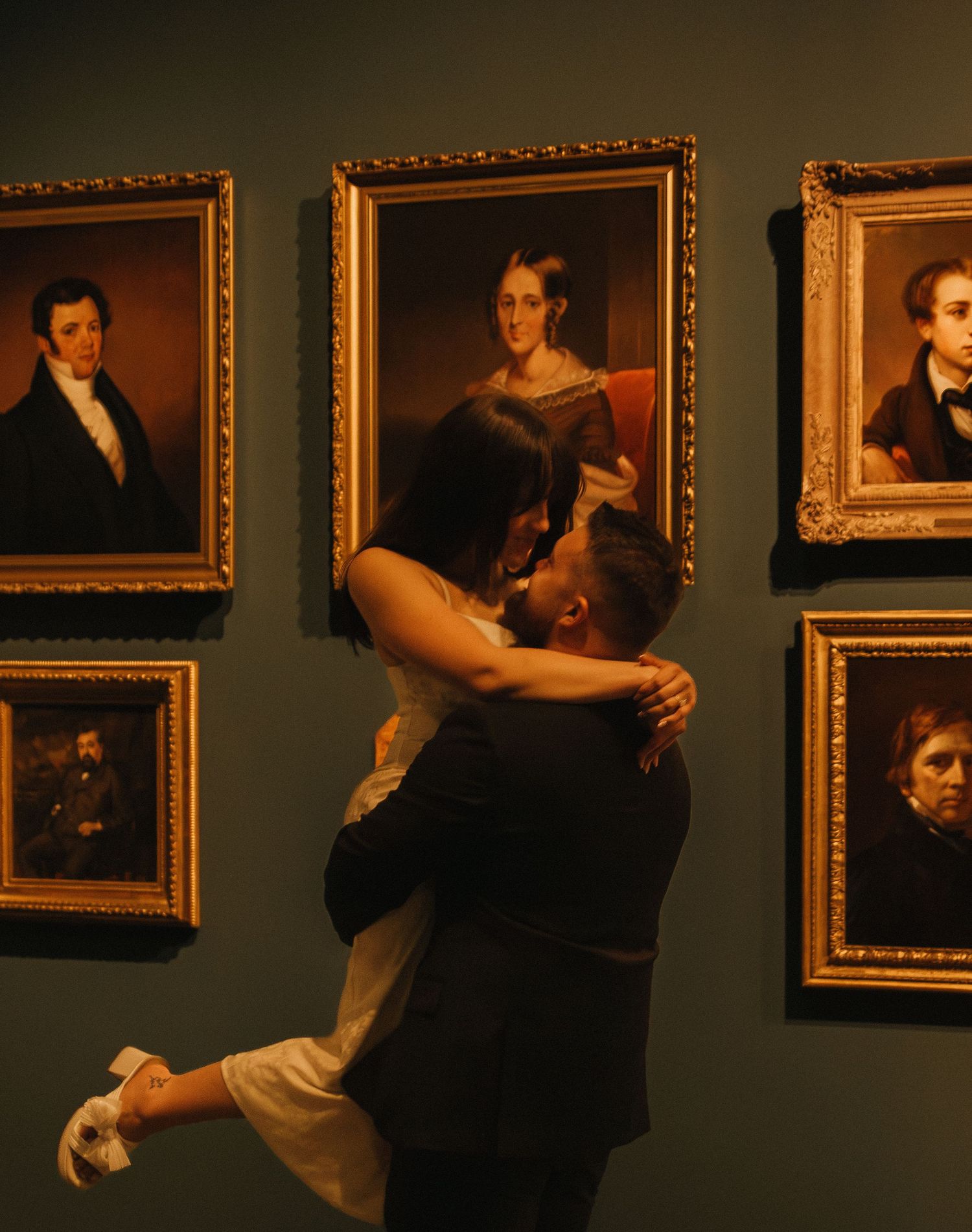 A couple dancing in an intimate embrace in front of a wall of classic portrait paintings in ornate gold frames.