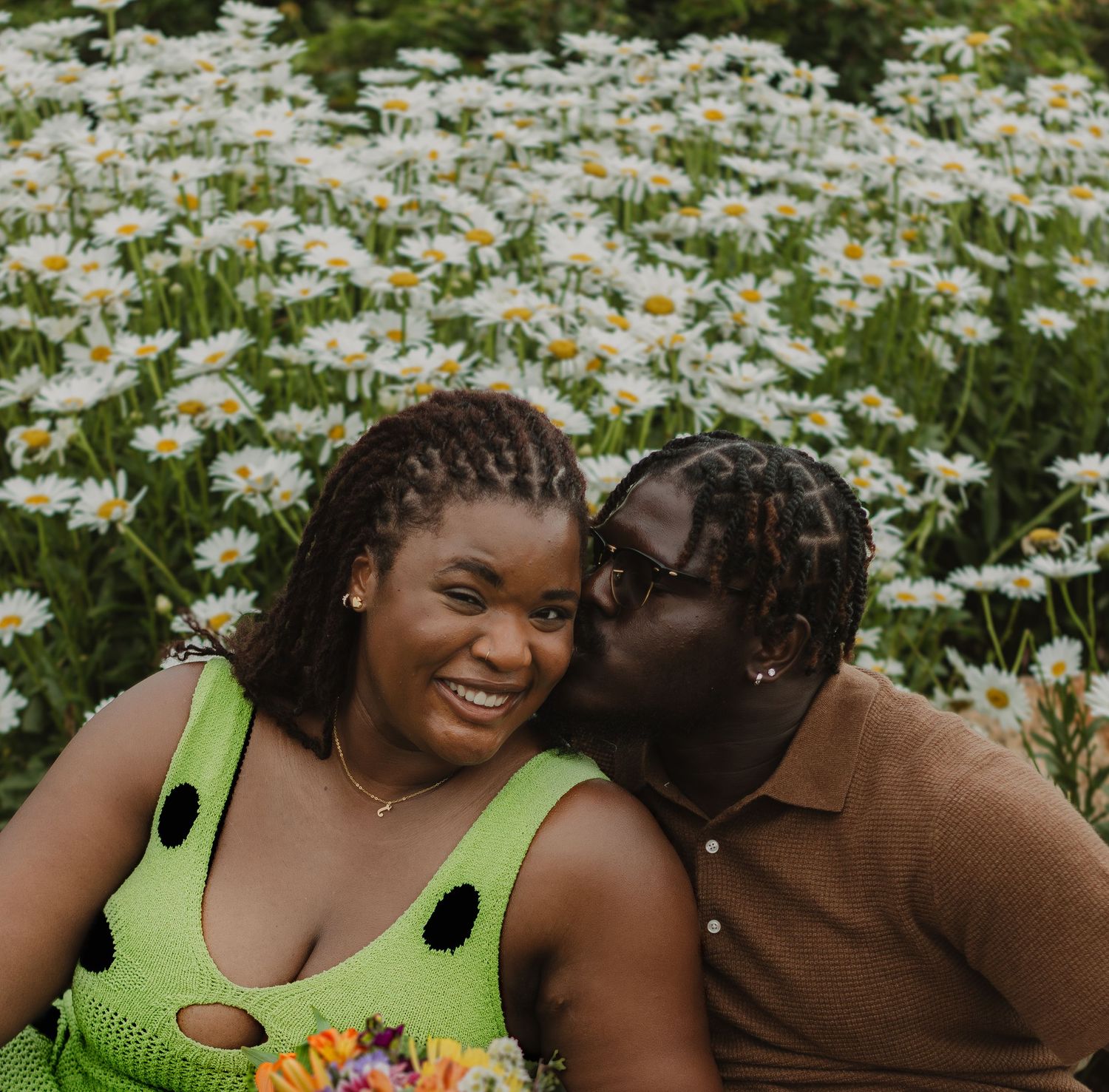 A couple shares a tender moment while sitting among blooming daisies in a garden.
