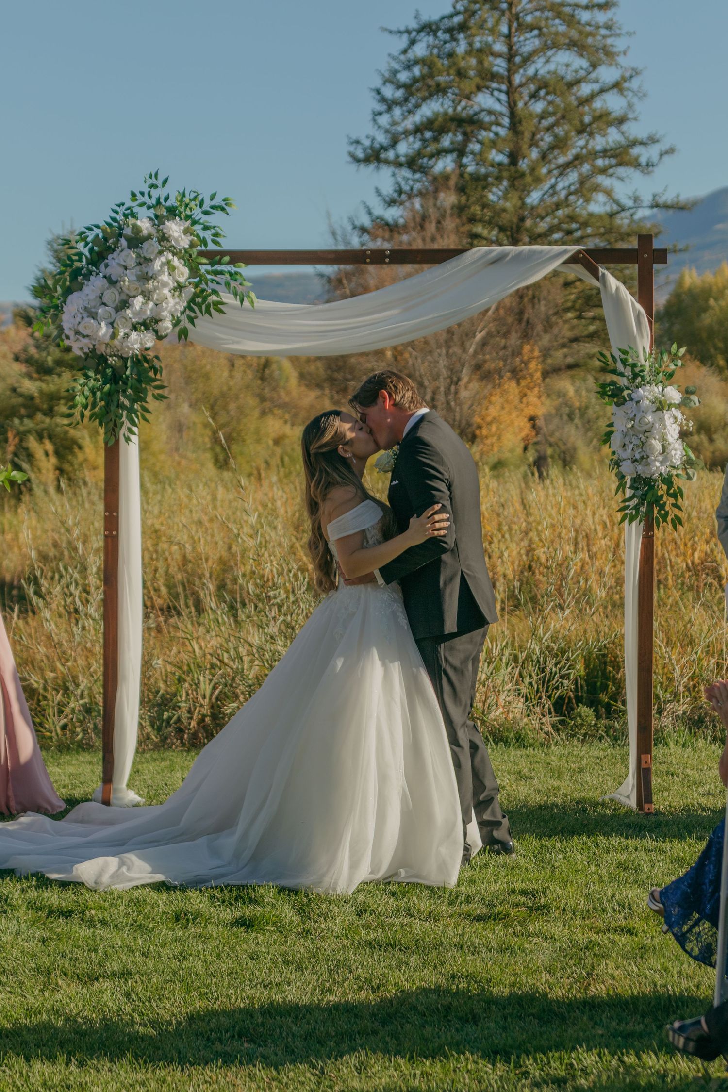 A romantic outdoor wedding ceremony with a white draped arch and floral decorations in a golden autumn field.