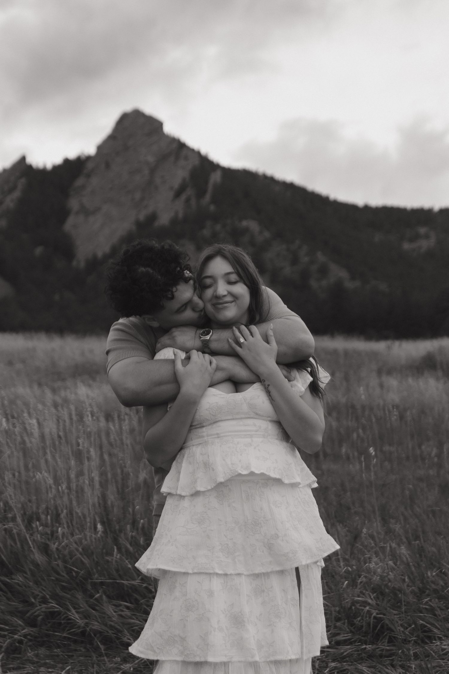Black and white romantic photo of man embracing woman from behind in field with mountain backdrop.