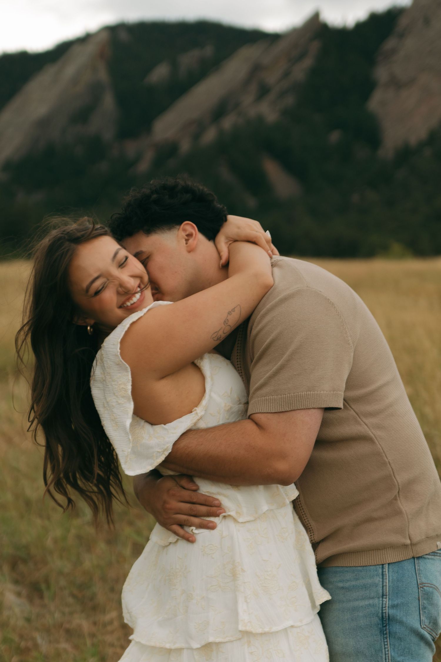A man embraces and kisses a smiling woman in a white dress in a scenic mountain meadow.