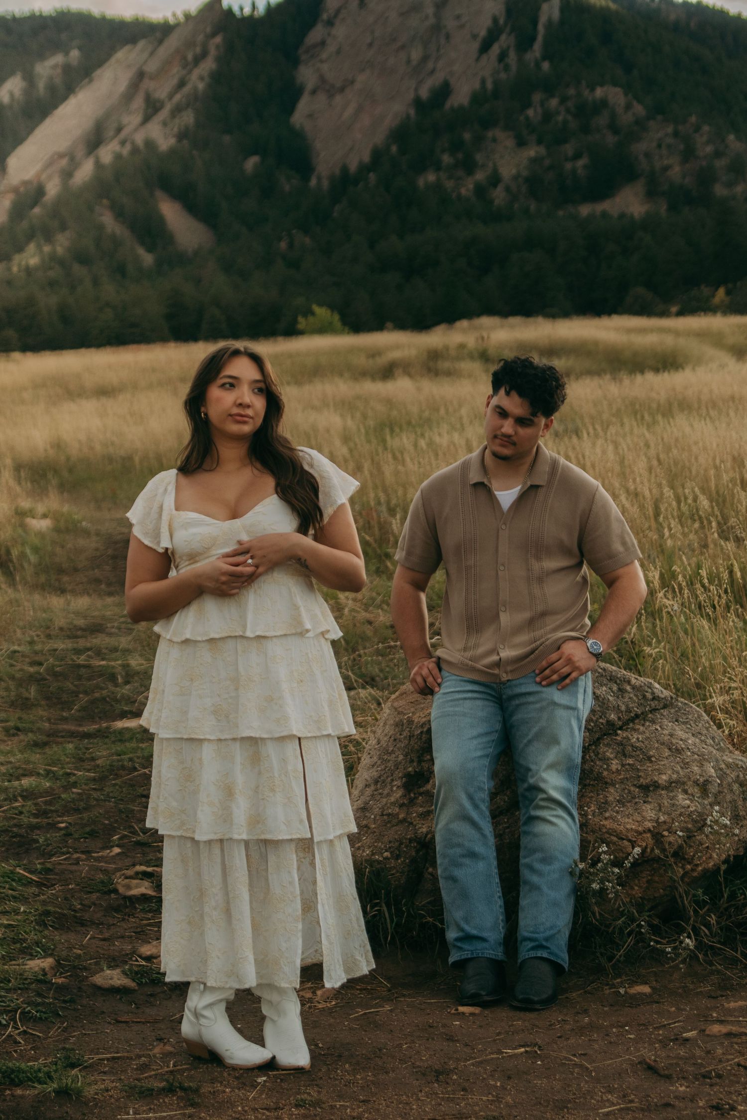Couple posing by boulder in golden field with dramatic mountain backdrop during engagement shoot.