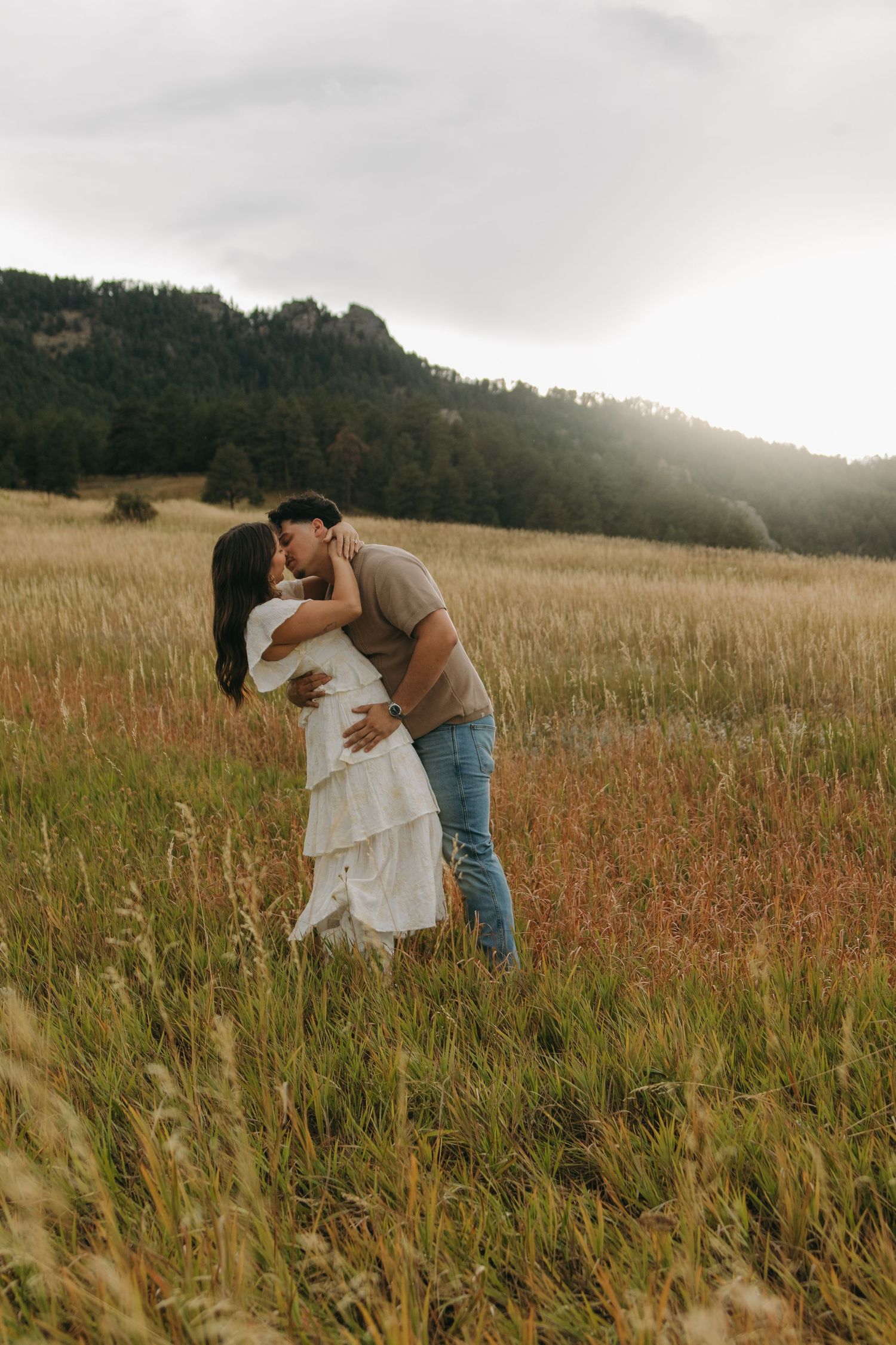 Couple sharing a romantic kiss in a golden meadow with mountain backdrop during an outdoor engagement session.