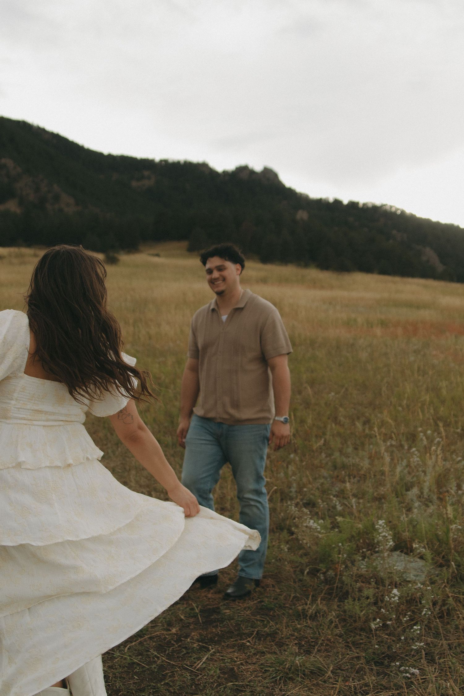 Woman in white dress twirls toward smiling man in tan shirt during romantic field engagement shoot.