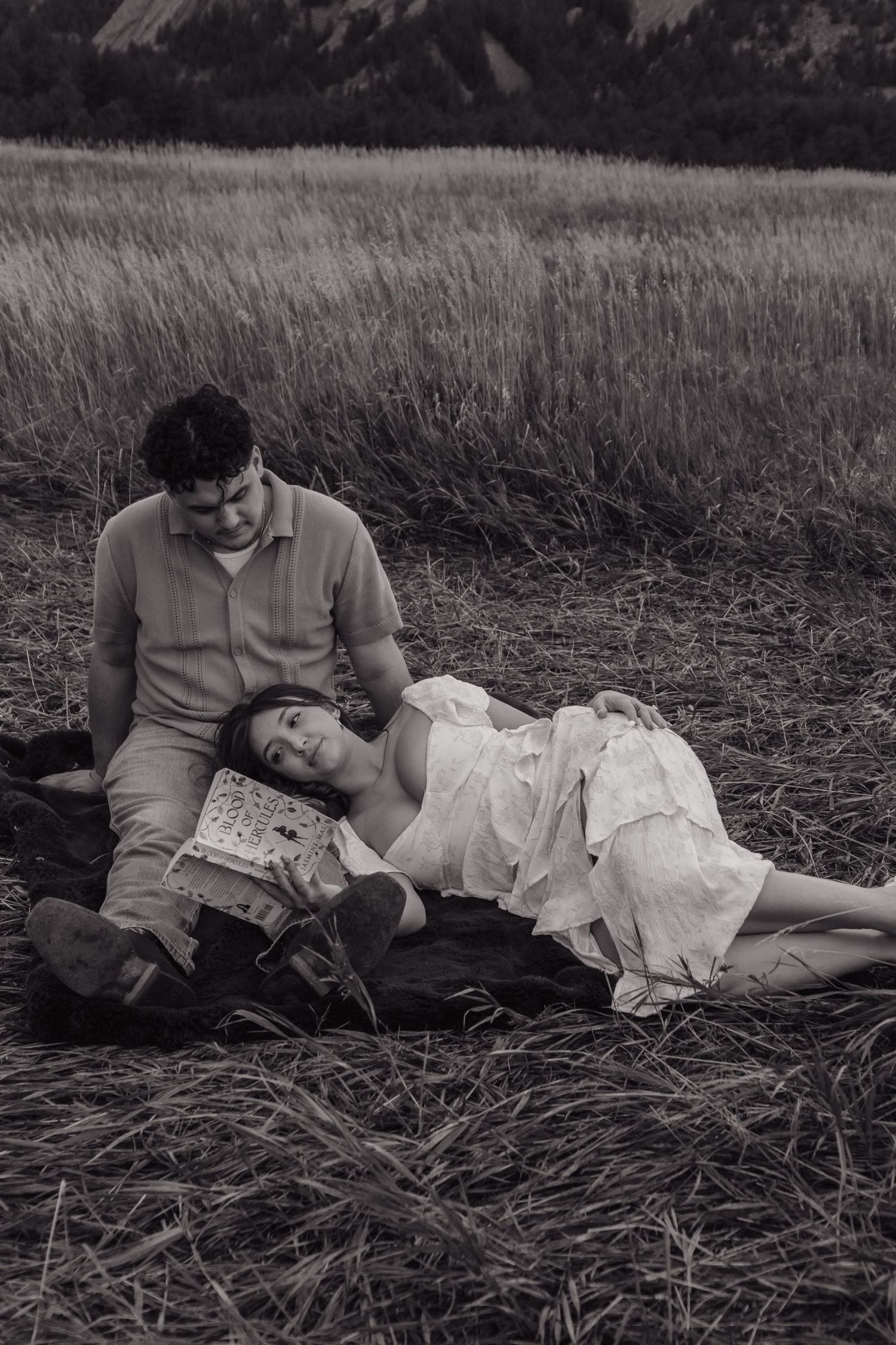 Black and white photo of woman lying in mans lap reading a book in a meadow.