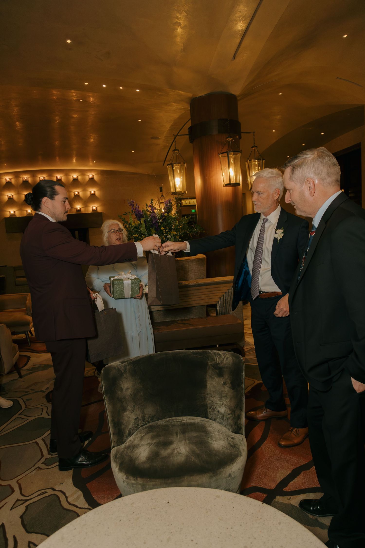 Groom in brown suit greeting guests with fist bump in warmly lit hotel lounge.