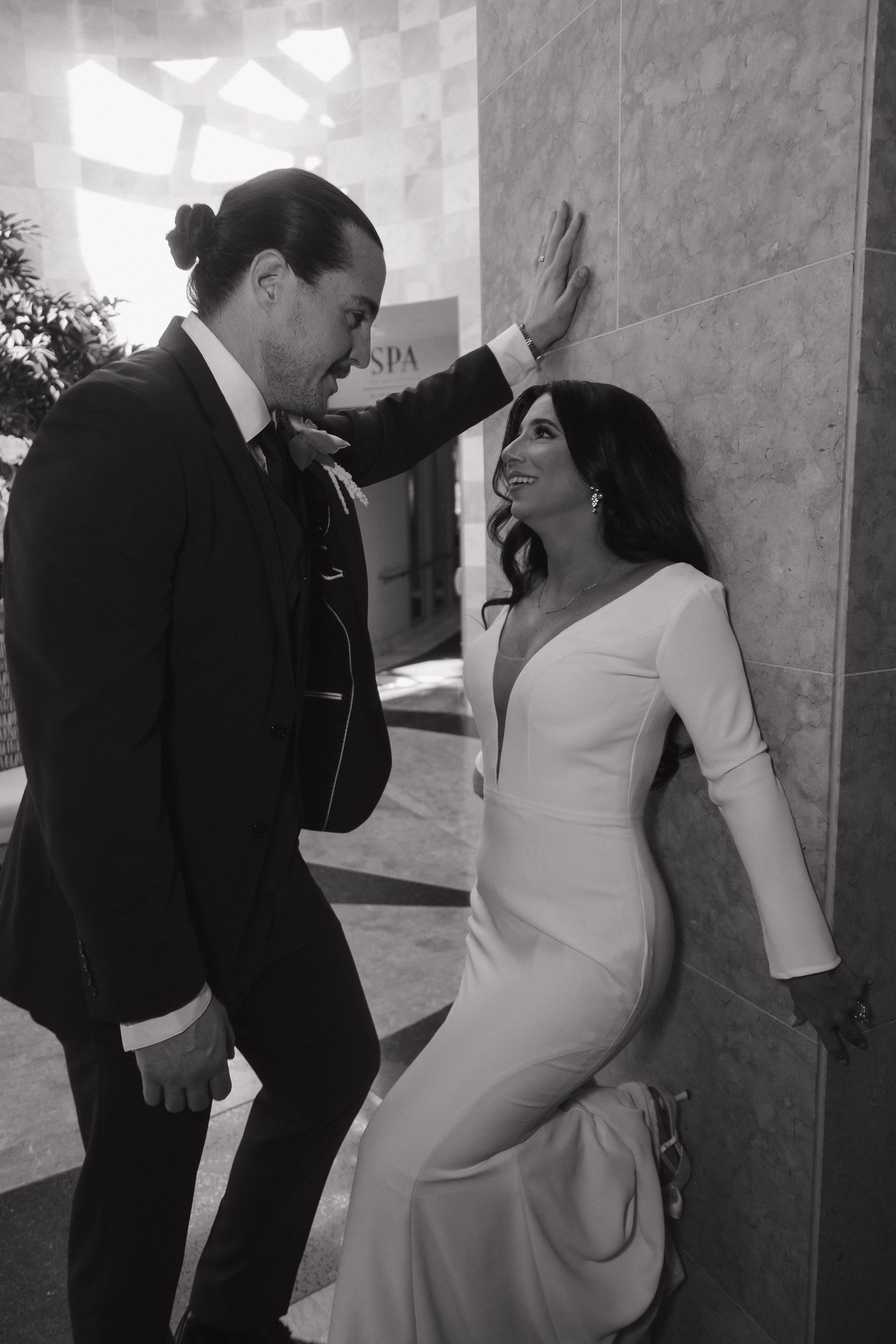 Black and white photo of groom leaning over bride against marble wall in elegant wedding attire.