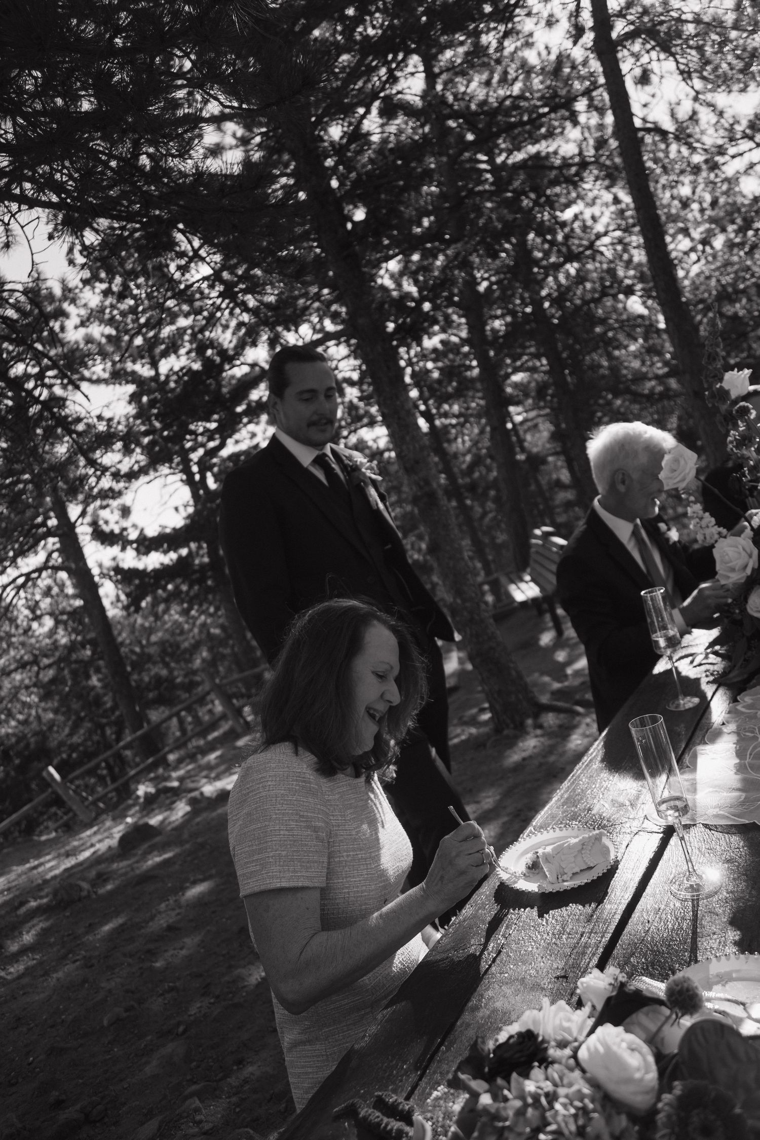 Black and white photo of wedding guests gathered at outdoor picnic table reception in pine forest.