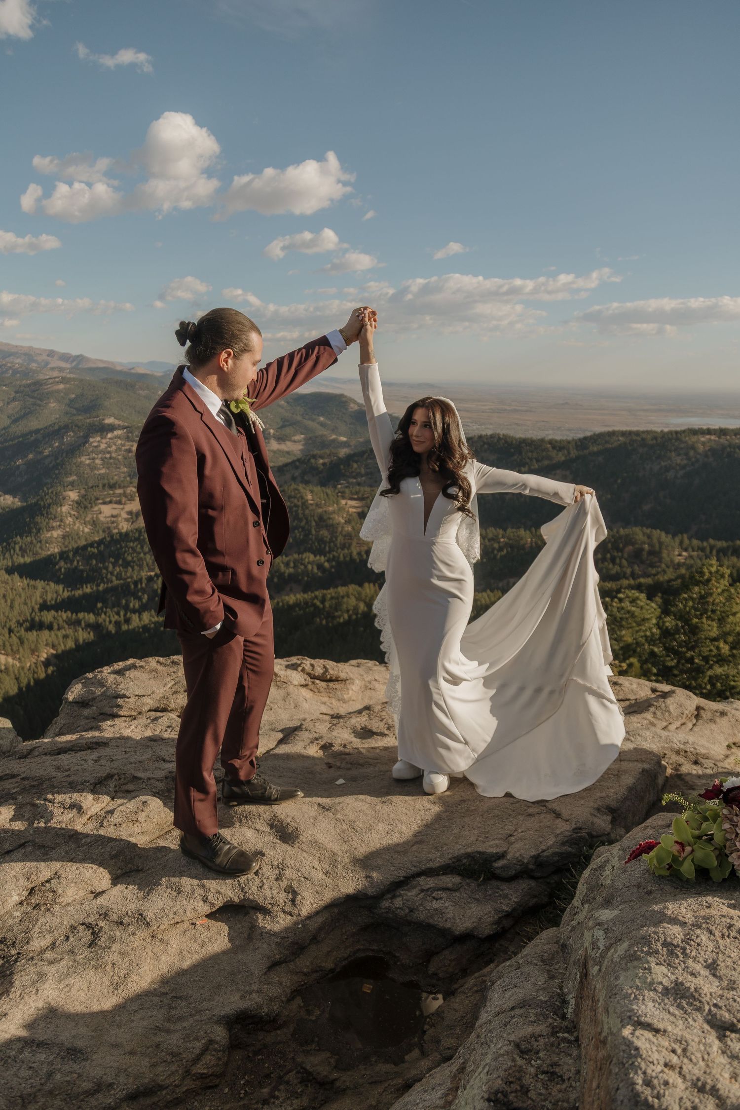 Bride and groom dance on a mountain summit, her white gown flowing in the wind against a scenic valley backdrop.