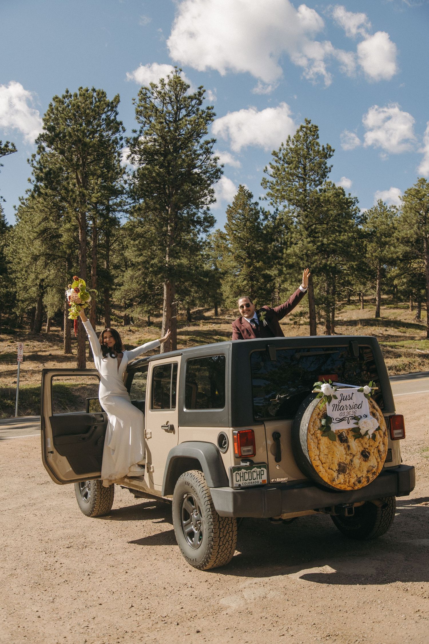 Joyful bride and groom celebrating beside a decorated Jeep Wrangler in a pine forest setting.