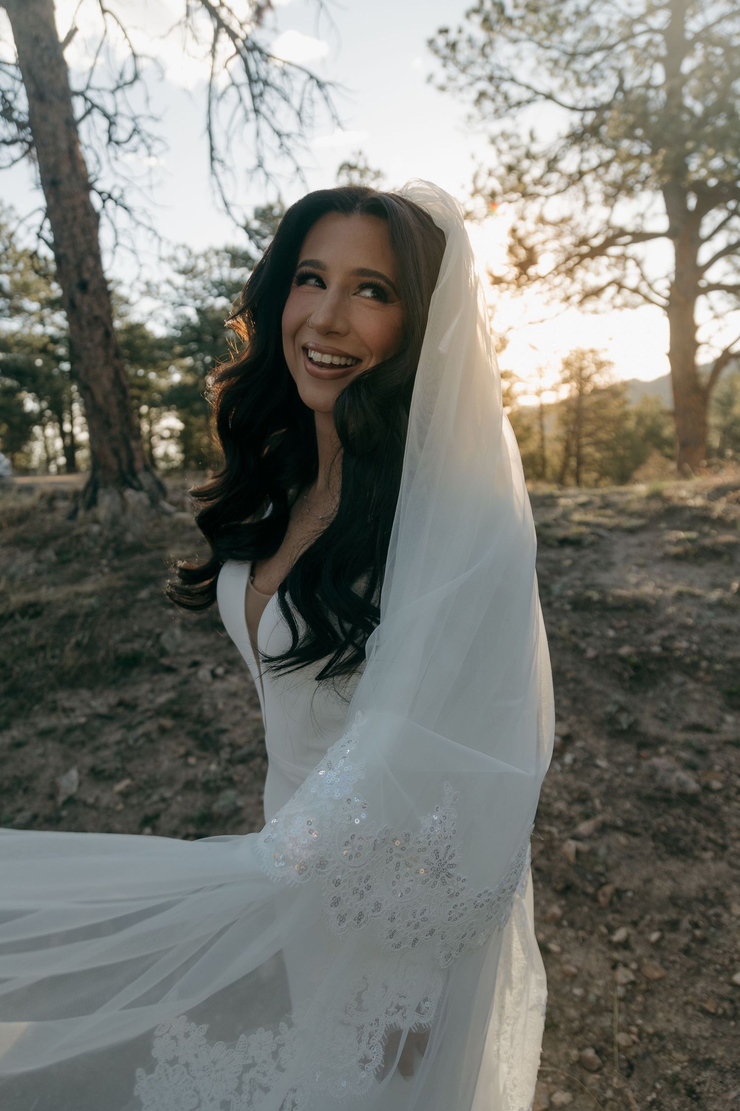 Radiant bride in white lace gown and veil smiles in sunlit forest wedding portrait.