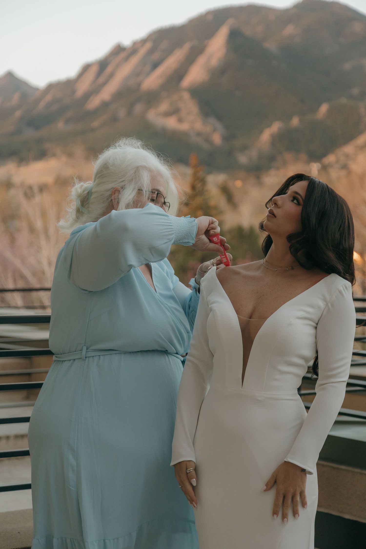 Older woman in blue dress helps bride with wedding gown details against stunning mountain backdrop.