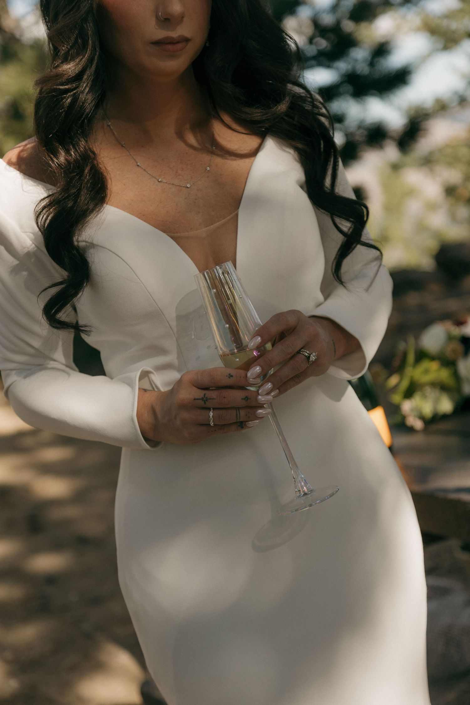 Bride in a white long-sleeve gown holding a champagne flute, showcasing her rings and delicate necklace.