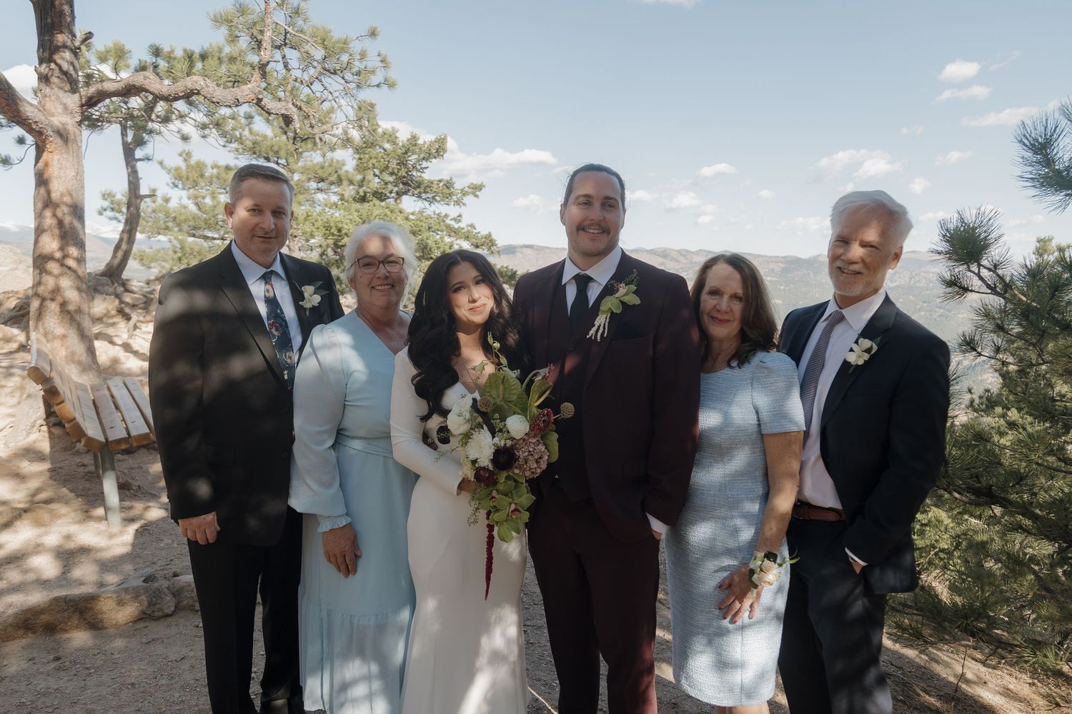 Wedding party poses outdoors on rocky overlook surrounded by pine trees under blue sky.