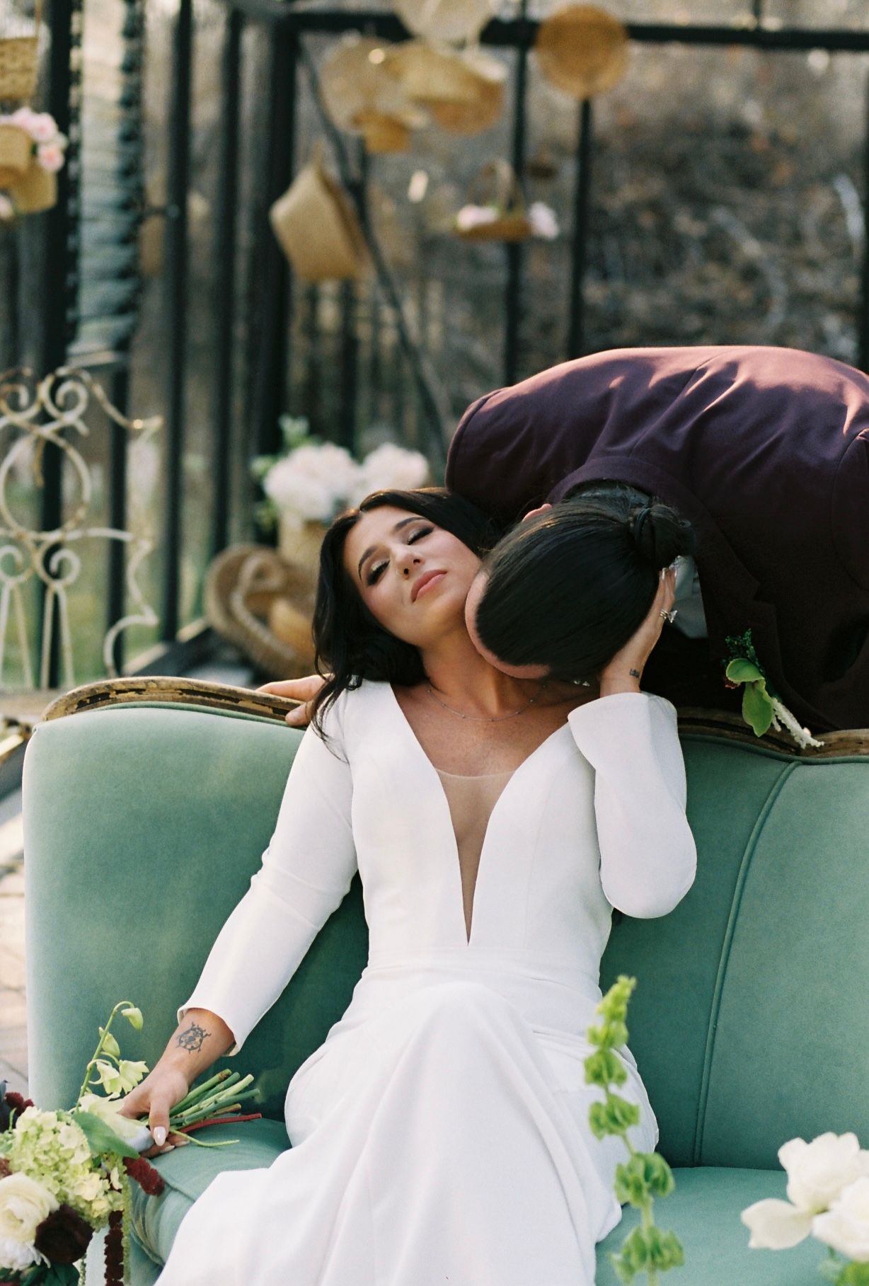 Bride in a deep V-neck white gown reclines on a teal sofa as groom leans down to kiss her cheek.