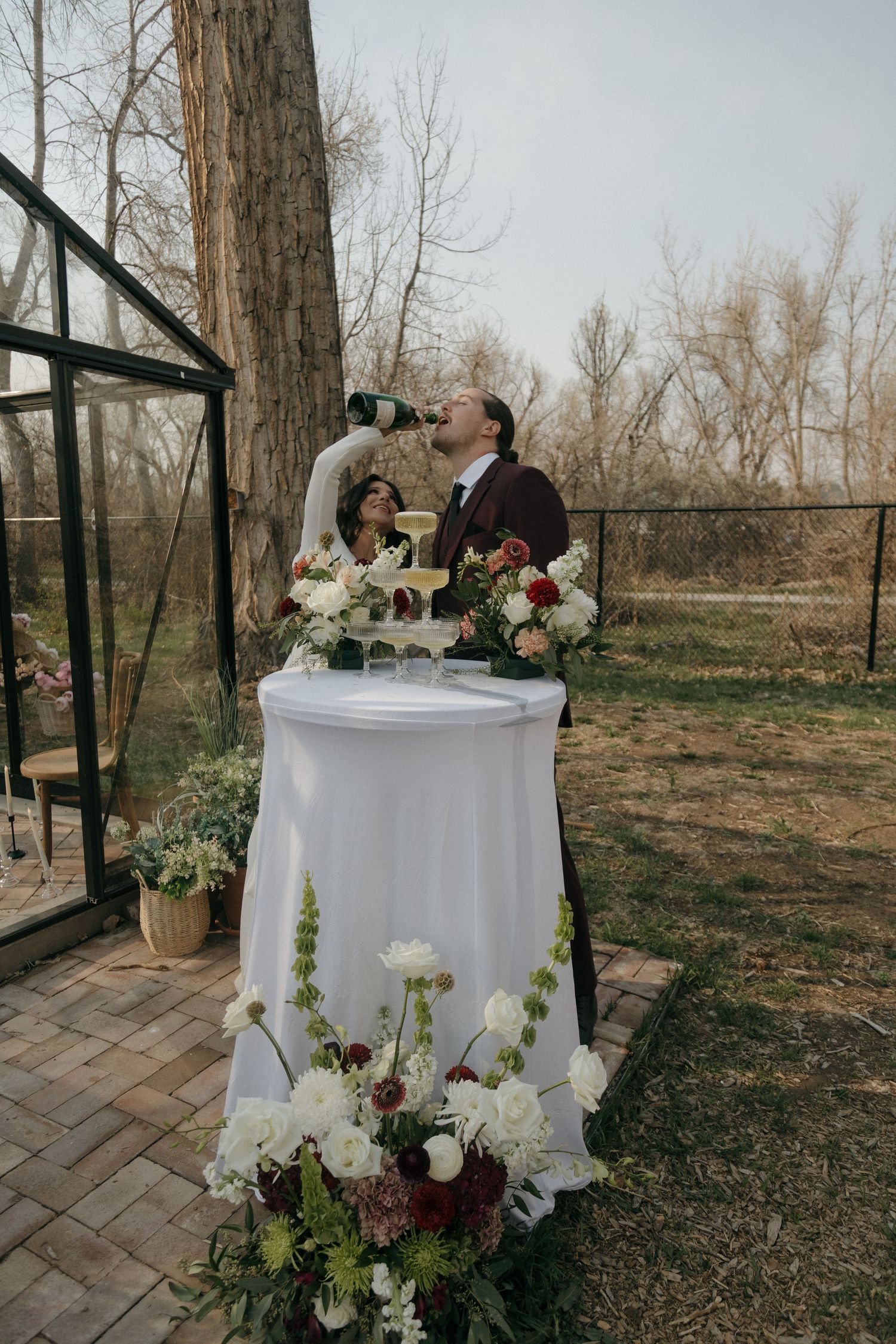 Bride and groom playfully drinking champagne directly from the bottle at their outdoor garden wedding.