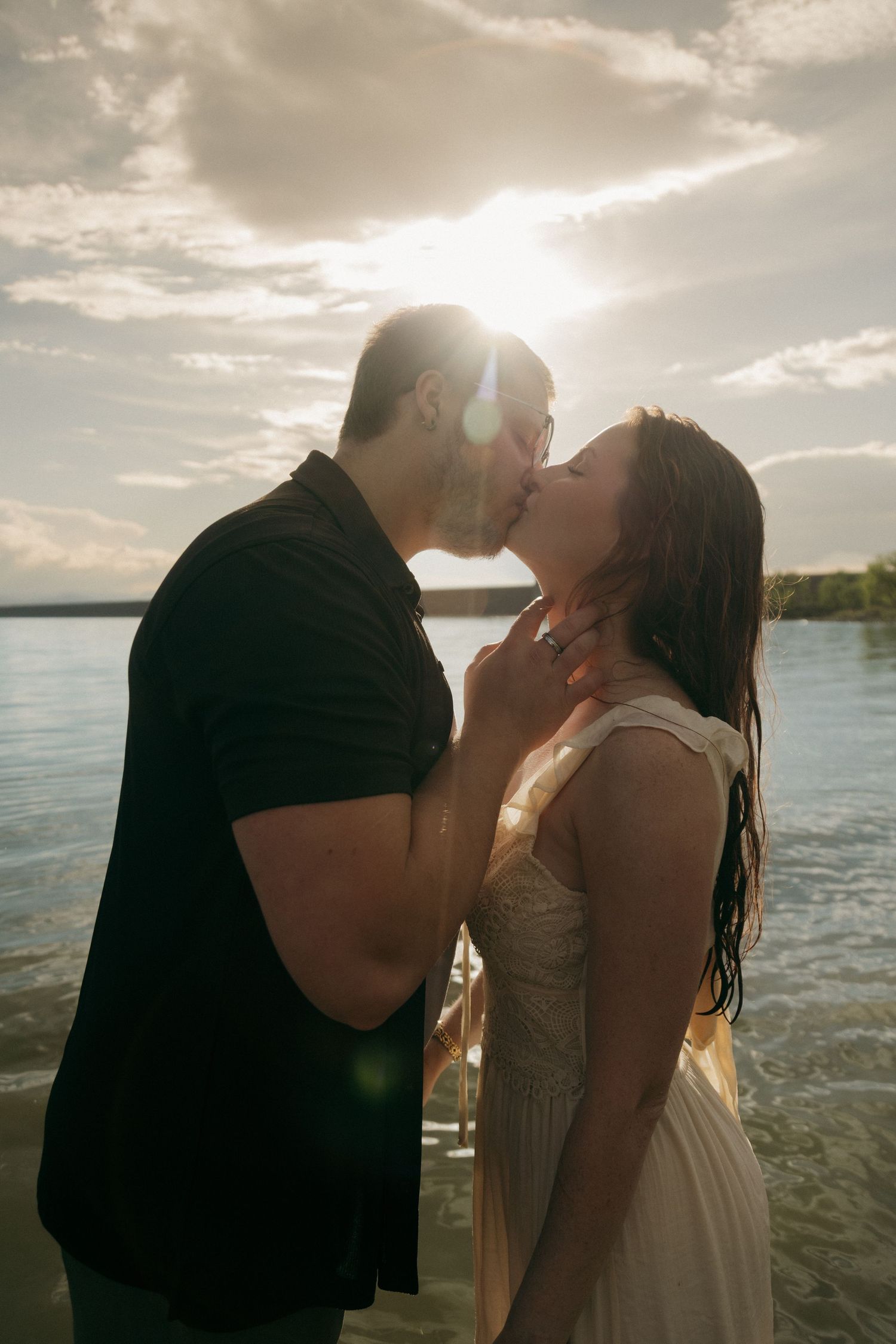 Couple kissing on lake shore backlit by golden sunset with lens flare effect.