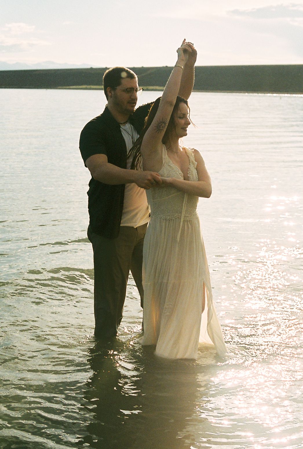Groom spinning bride in white dress in shallow lake water, holding her hand raised under bright sunlight.