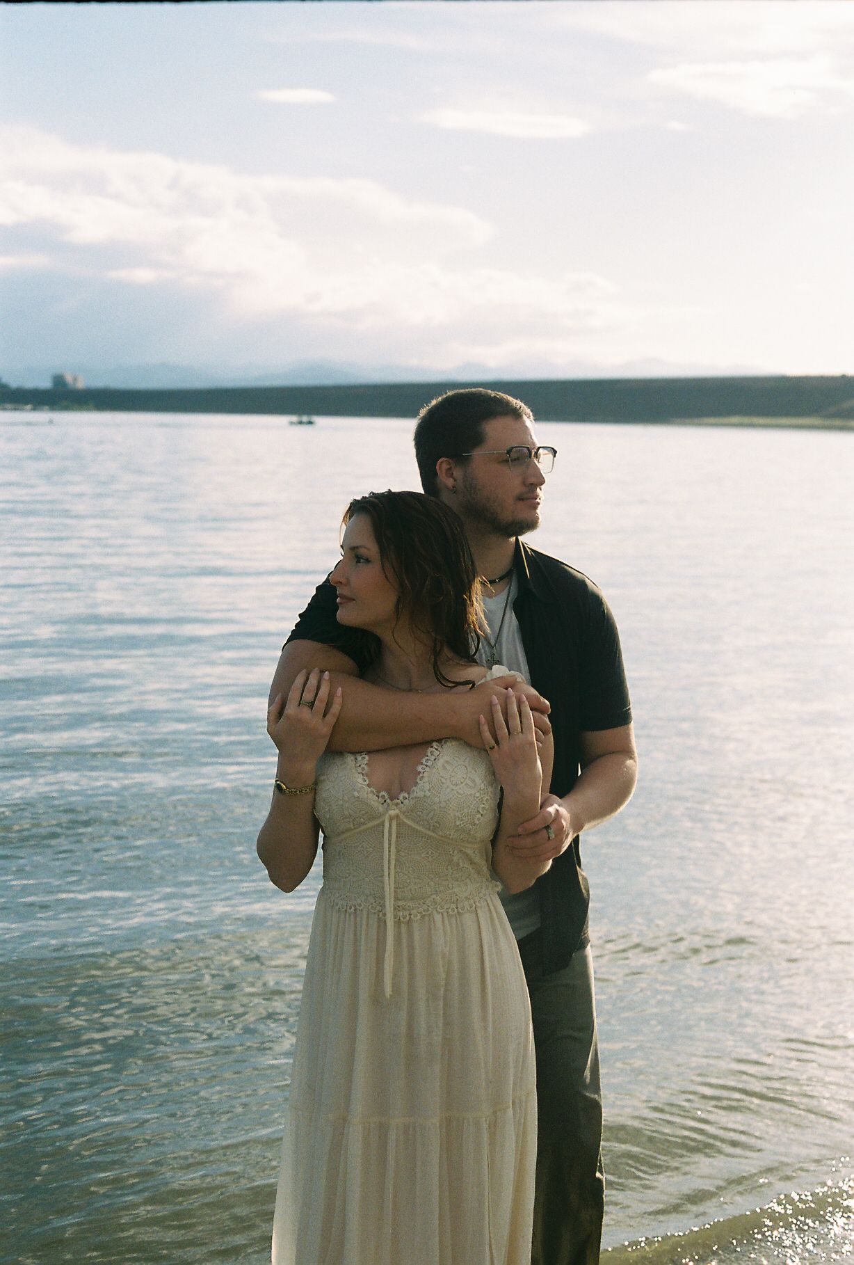Couple embracing on lake shore at sunset, woman in white lace dress, man in black.