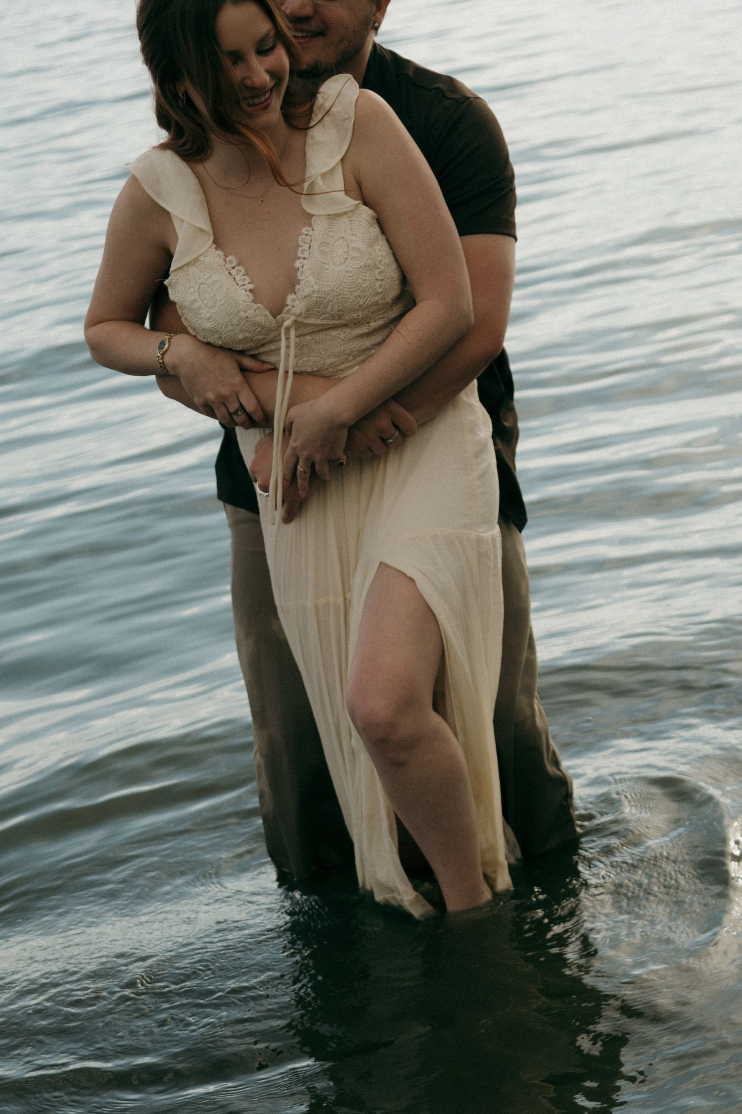 Couple embracing in water, woman in elegant white lace dress with slit, romantic outdoor engagement photo session.