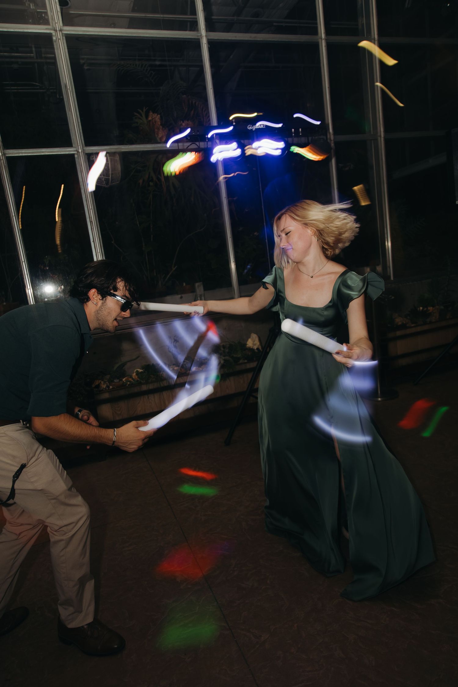 Wedding guests duel playfully with glowing light wands on colorful dance floor at reception.